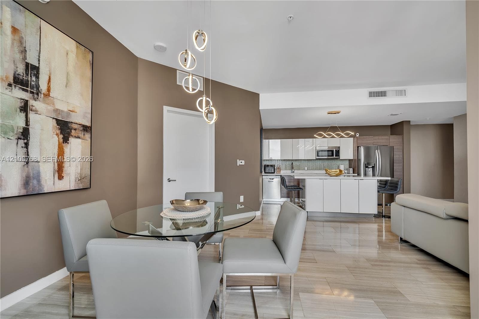 Open-concept interior featuring large-format tile flooring, modern multi-tier LED pendant lighting, white flat-panel kitchen cabinetry, a kitchen island with stone-surface countertop, and stainless steel appliances