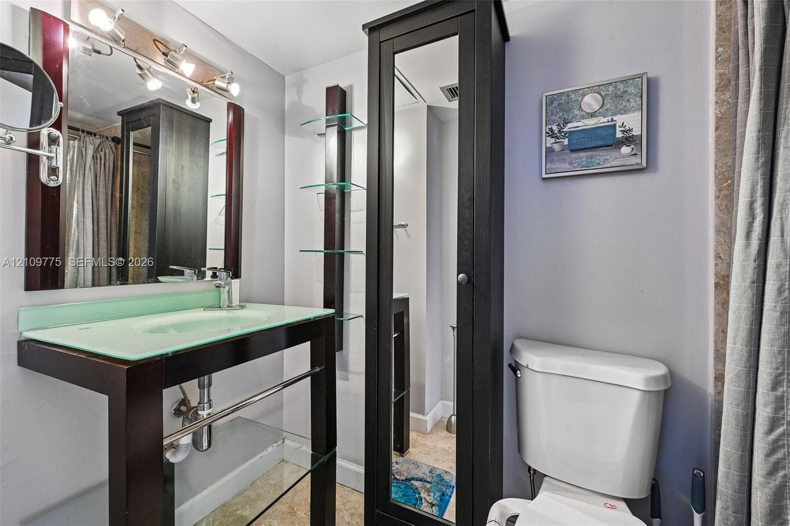 Bathroom featuring a floating vanity with a glass countertop, integrated sink, polished metal hardware, a tall mirrored storage cabinet, and built-in glass shelving