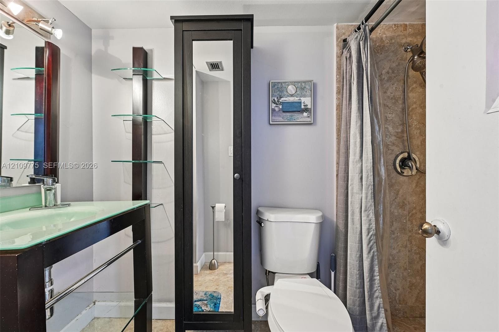 Bathroom featuring a glass-topped vanity with an integrated basin, a tall dark-finish built-in storage cabinet with a mirrored door, glass shelving, and a tiled shower stall with a hand-held shower head