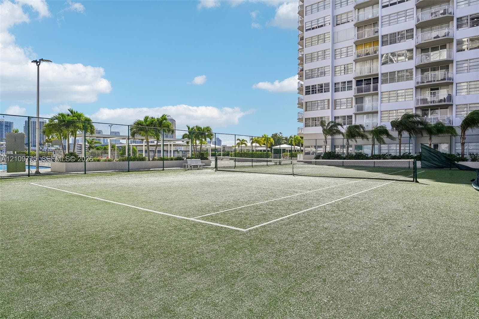 Tennis court with synthetic turf surface, enclosed by chain-link fencing, featuring overhead lighting and perimeter landscaping with palm trees