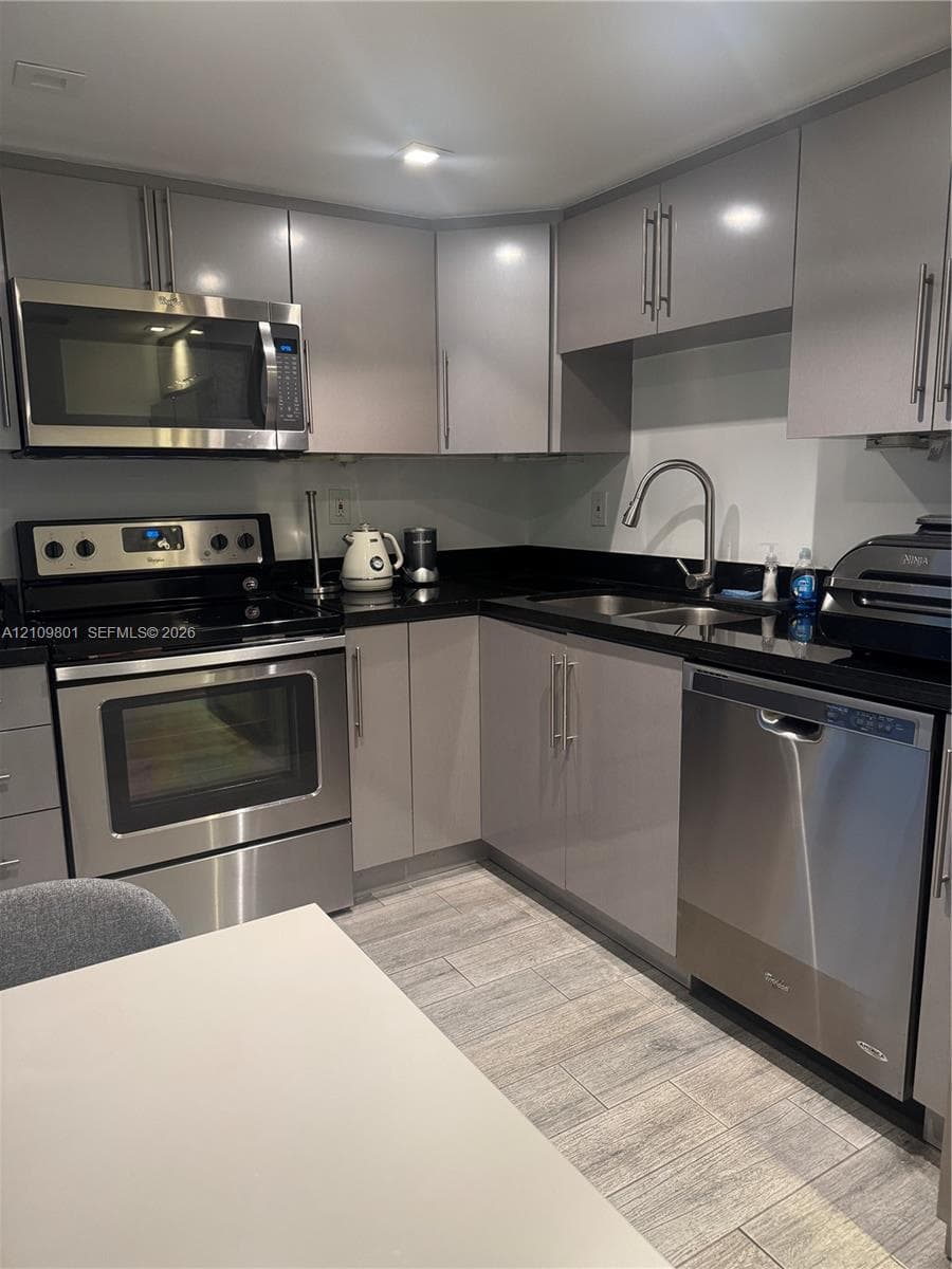 Kitchen featuring grey flat-panel cabinetry, black stone countertops, stainless steel appliances, a dual-basin sink with a gooseneck faucet, and wood-finish tile flooring