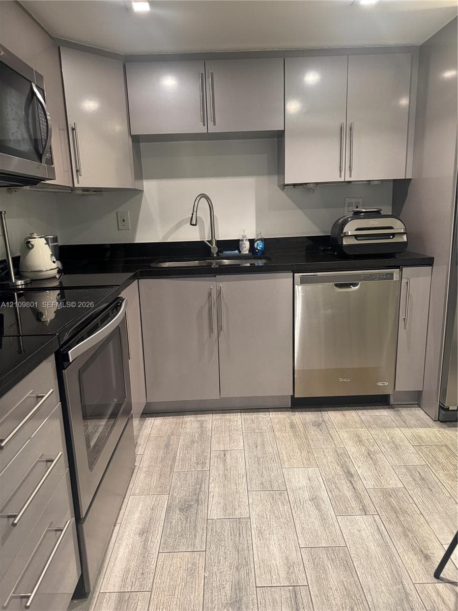 Kitchen featuring gray slab-front cabinetry, black stone countertops, wood-finish tile flooring, and stainless steel appliances including a built-in dishwasher and microwave