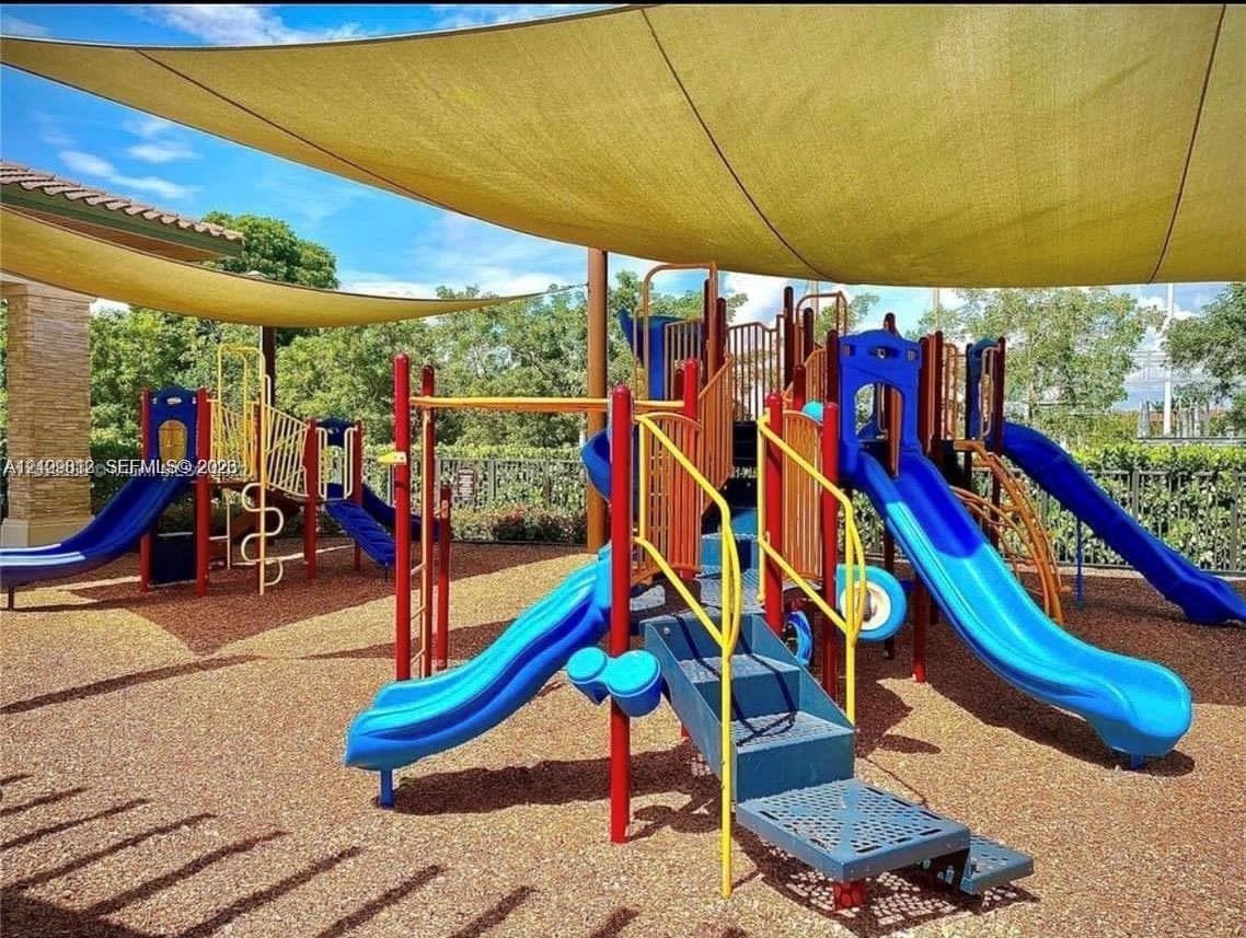 Outdoor recreational area featuring a multi-level climbing structure with integrated slides, soft-surface ground cover, and overhead shade canopies enclosed by perimeter metal fencing