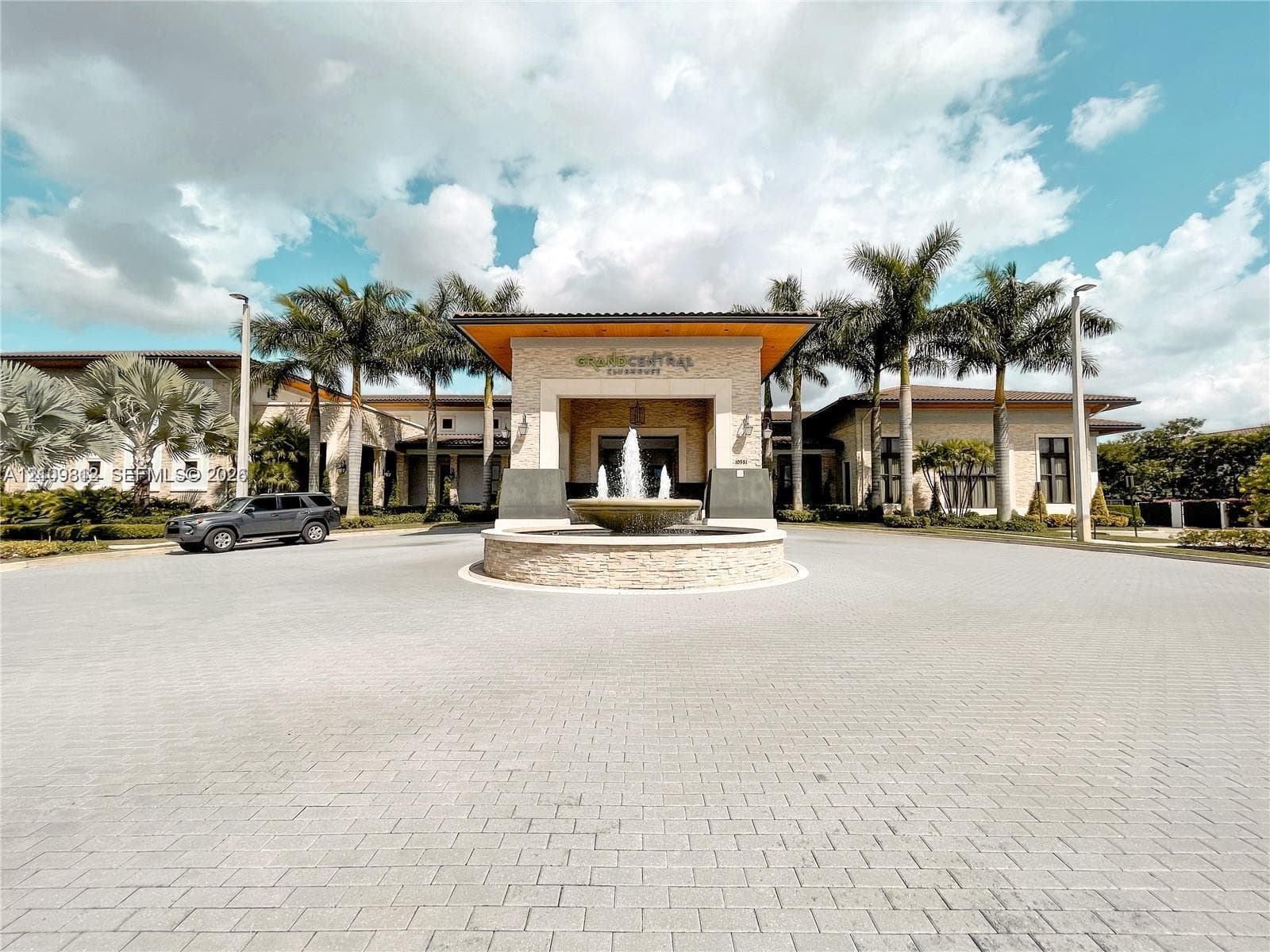 Clubhouse exterior featuring a stone-clad portico, paved circular driveway, central water fountain, and mature palm landscaping