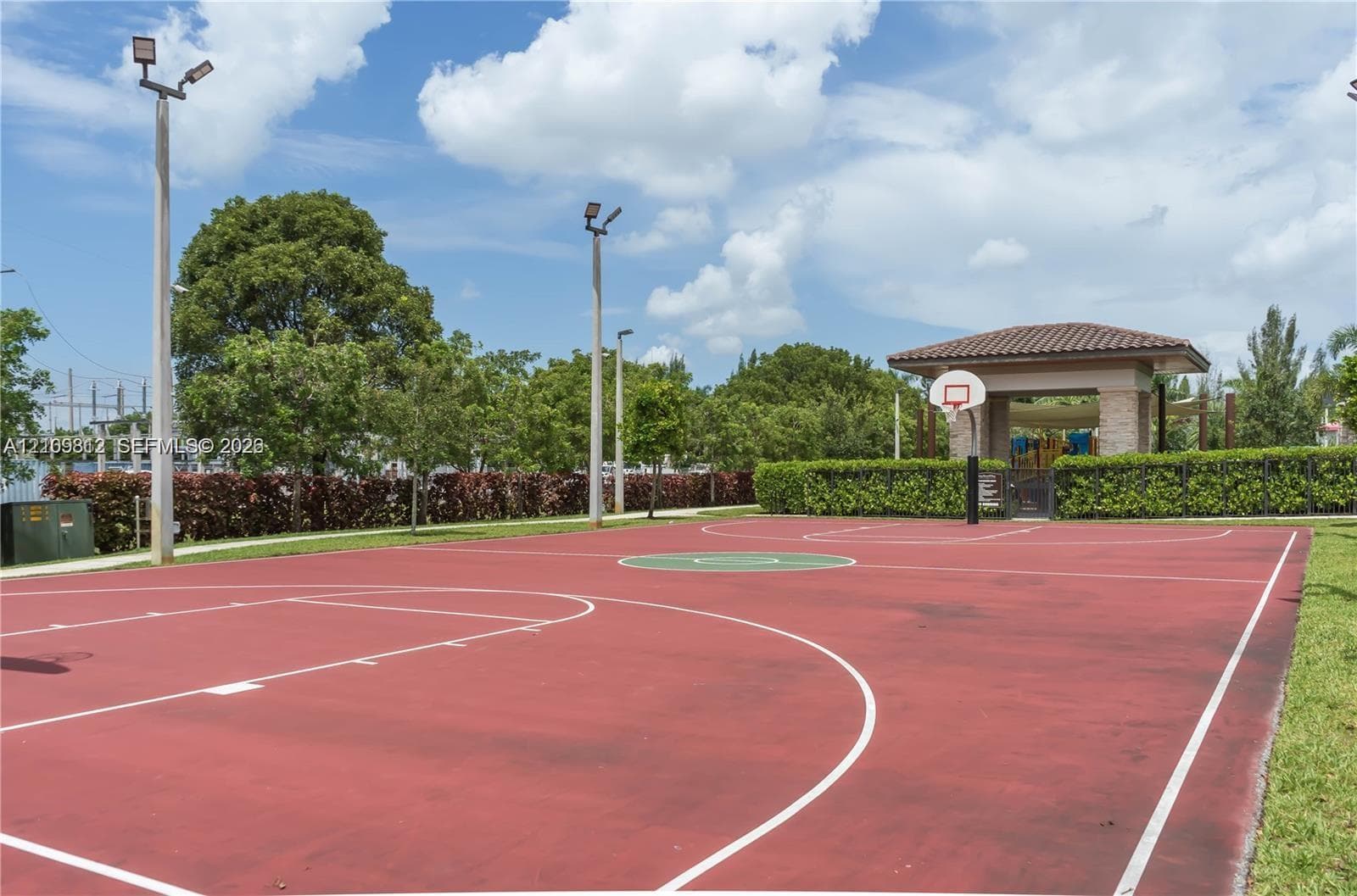 Outdoor basketball court featuring a red court surface, integrated lighting, and a roofed structure