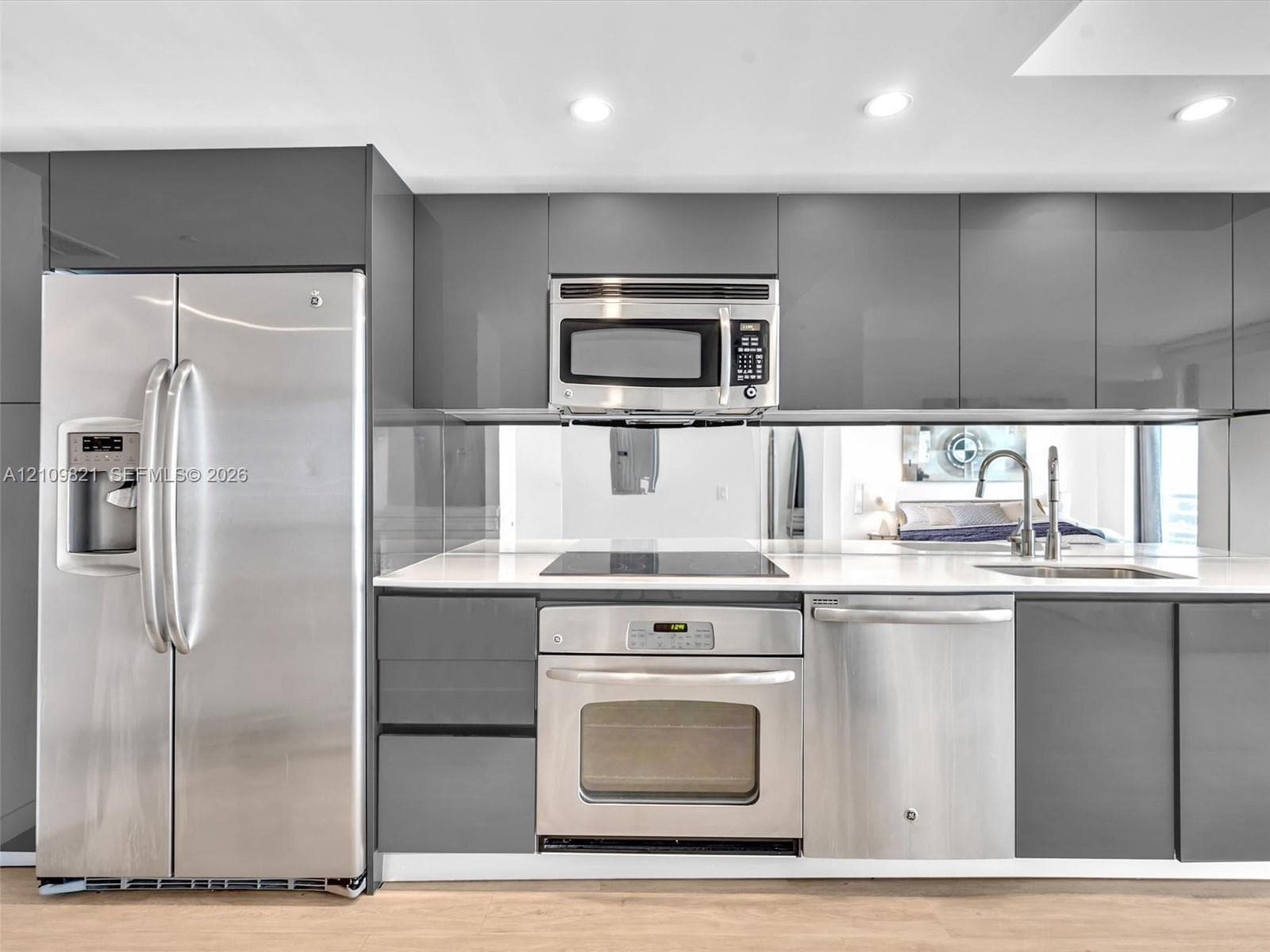 High-gloss grey cabinetry featuring sleek hardware, stainless steel appliances, a mirrored backsplash, integrated cooktop, and solid-surface countertops