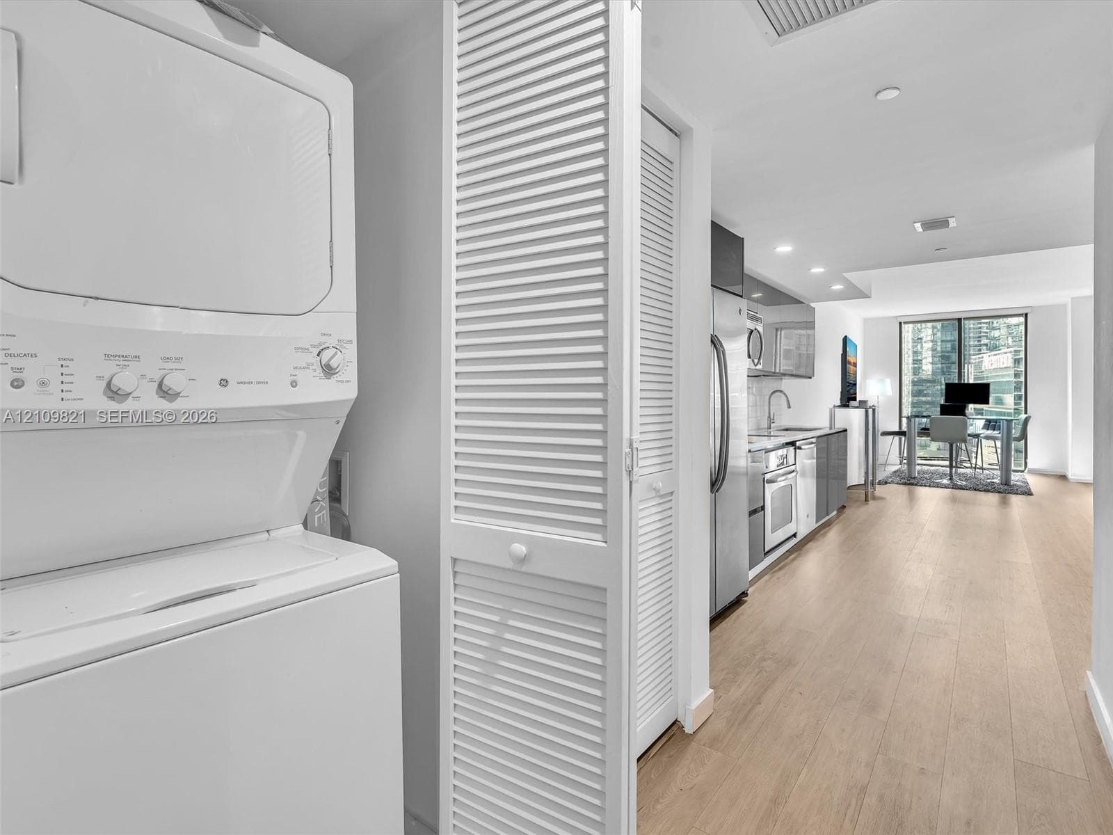 Stacked laundry unit enclosed by white louvered doors, light-toned wood-finish flooring, stainless steel kitchen appliances, and recessed ceiling lighting