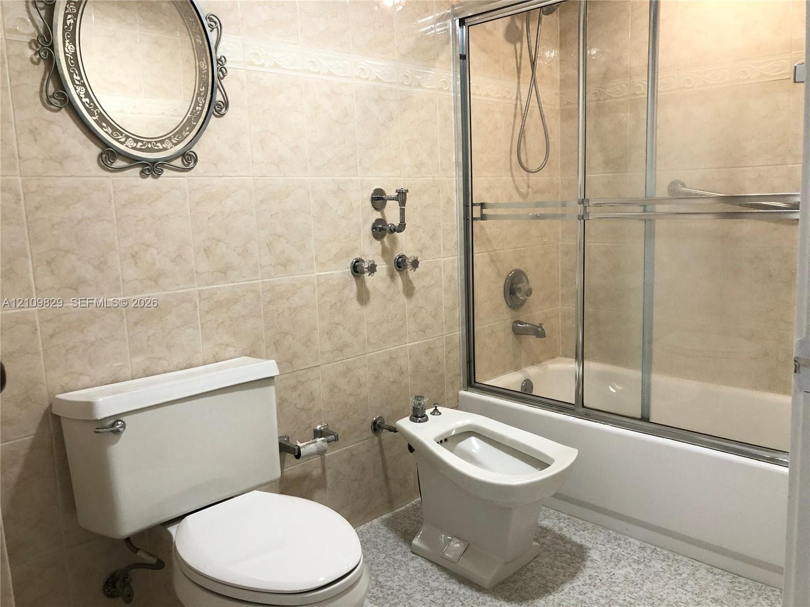 Bathroom featuring wall-to-wall ceramic tile, a soaking tub with a sliding glass enclosure, a pedestal bidet, and a wall-mounted metallic mirror