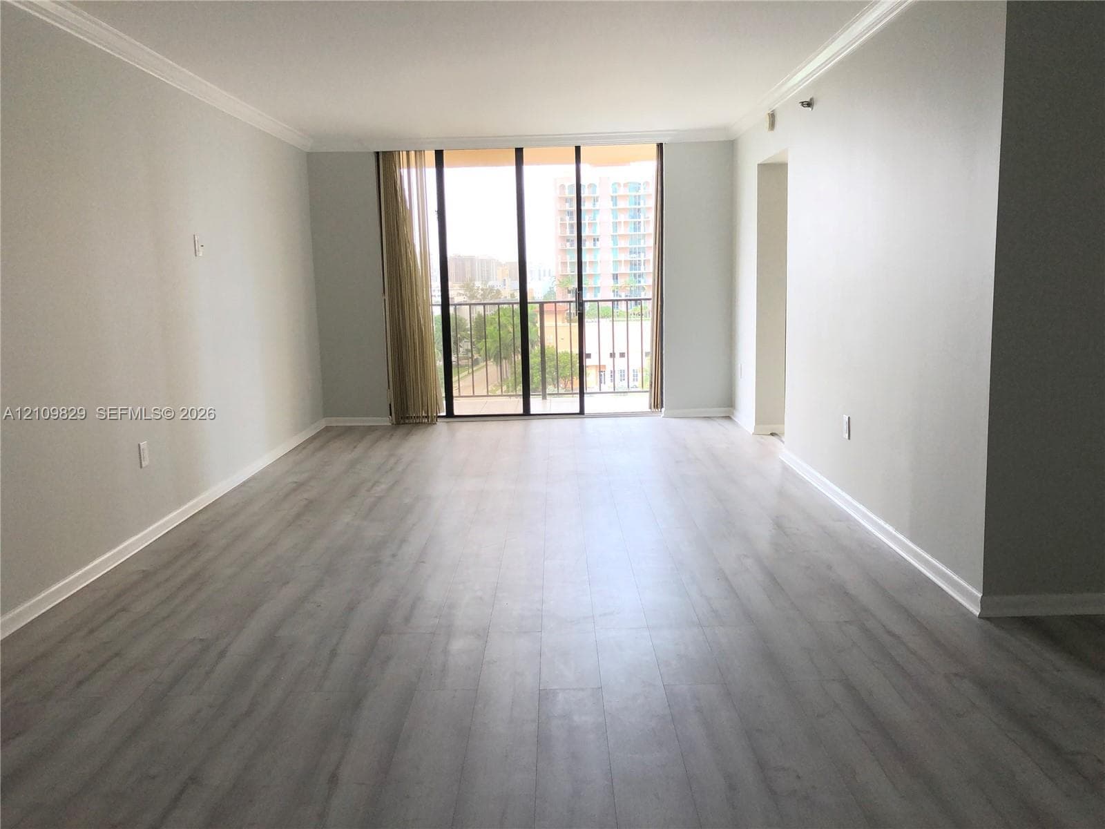 Open-concept room featuring light-toned wood-finish flooring, neutral wall paint, crown molding, and floor-to-ceiling glass sliding doors opening to a balcony