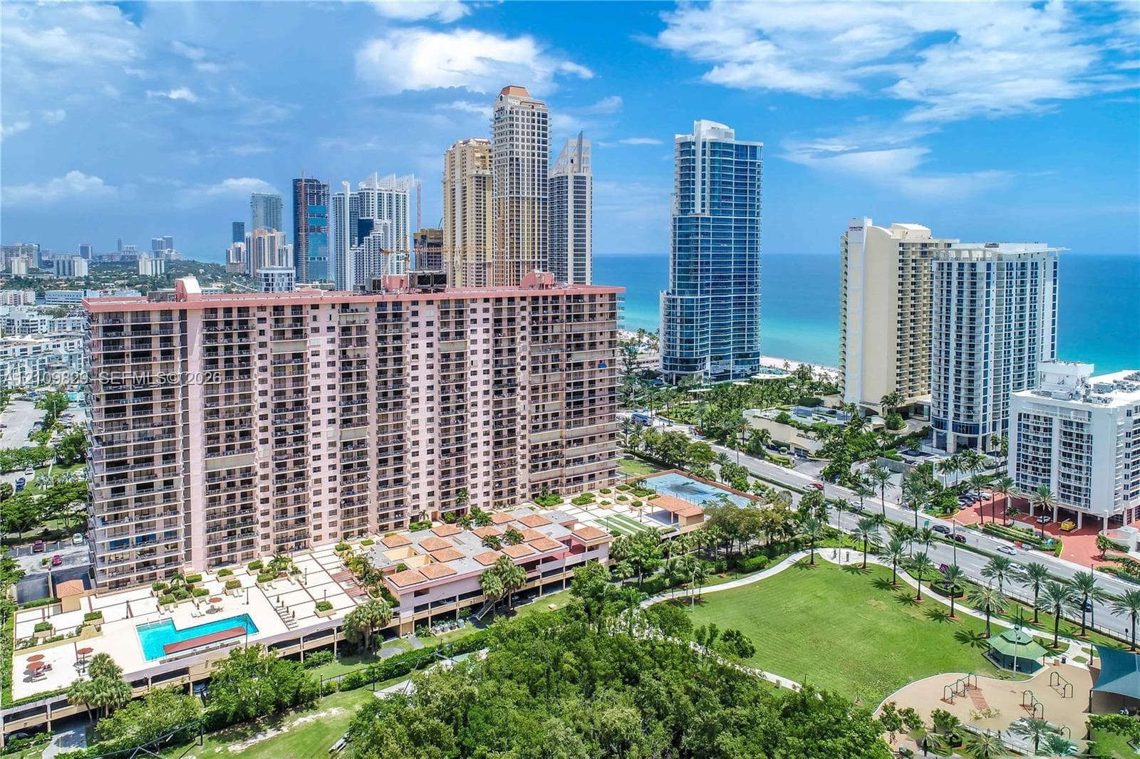 High-rise residential building featuring expansive private balconies, a ground-level swimming pool, multi-level parking structure, and proximity to municipal park grounds