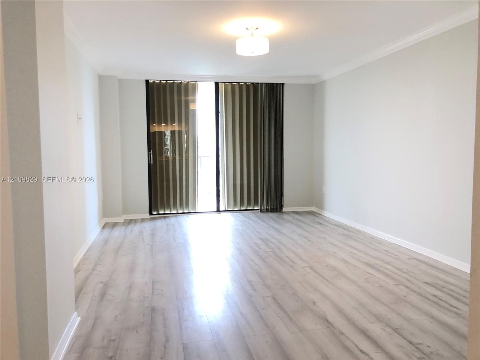 Living area featuring light-toned wood-finish flooring, crown molding, and sliding glass balcony doors with vertical blinds