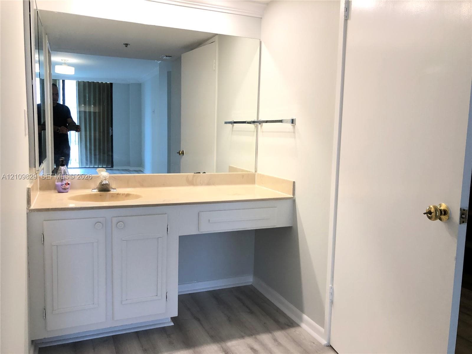 Vanity featuring a light-toned countertop with an integrated basin, white cabinetry with recessed panels, a full-width wall mirror, and wood-finish flooring