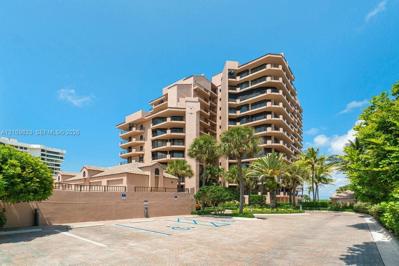 Multi-story high-rise building featuring private balconies, masonry exterior, paved parking area, and mature palm trees