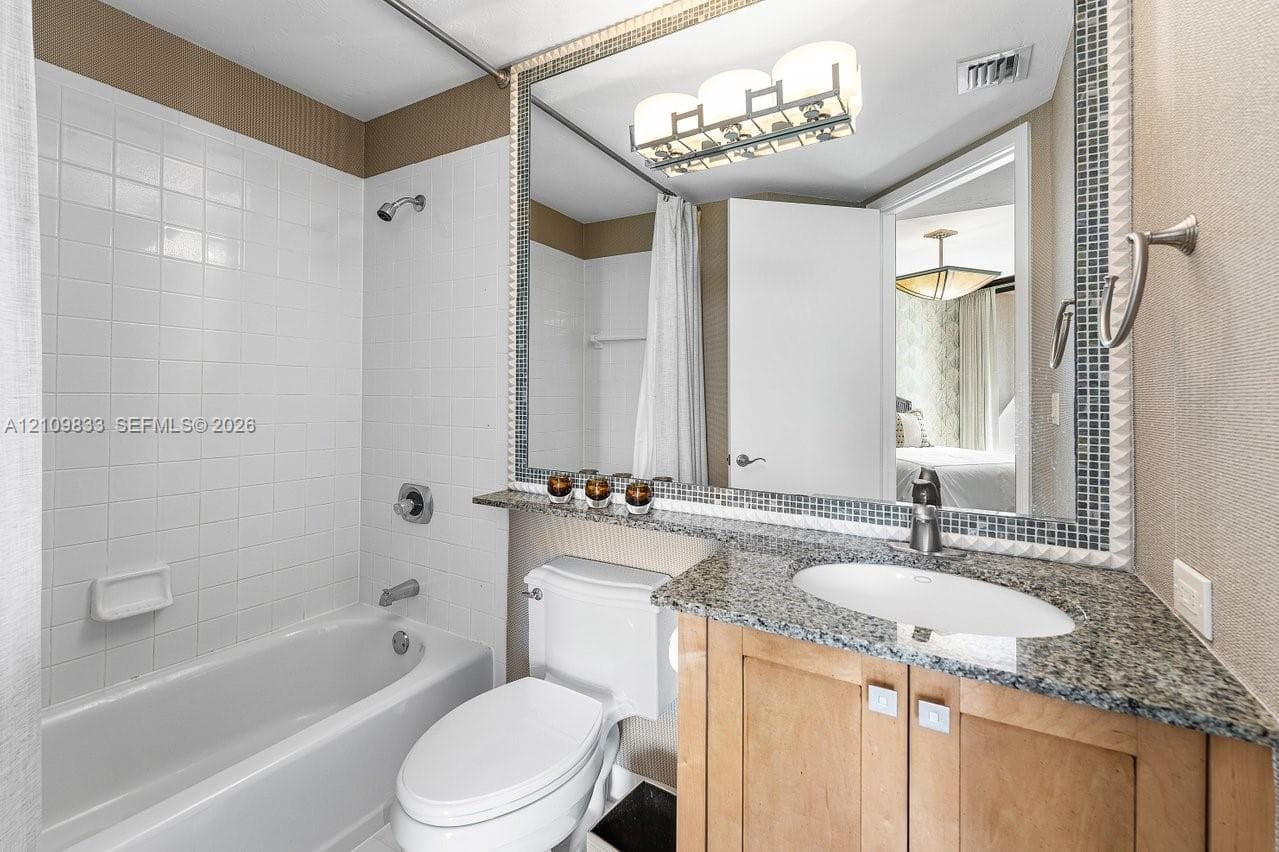Bathroom featuring a vanity with stone-finish countertop, undermount sink, large mirror with mosaic tile trim, white tile shower surround, and contemporary three-light fixture