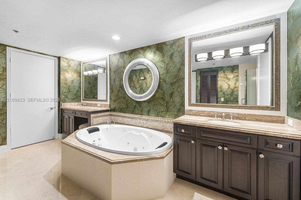 Oval jetted tub with tiled surround, dual dark-finish wood cabinetry, integrated lighting, stone-top vanities with undermount sinks, and wall-mounted mirrors
