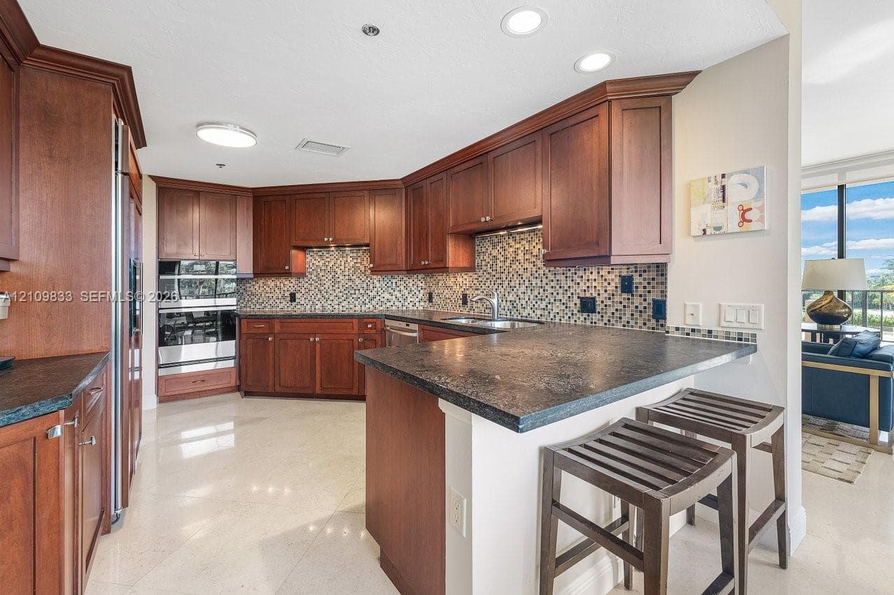 Kitchen featuring dark wood cabinetry, integrated stainless steel double wall ovens, a multi-tone tile backsplash, dark stone countertops, and a breakfast bar