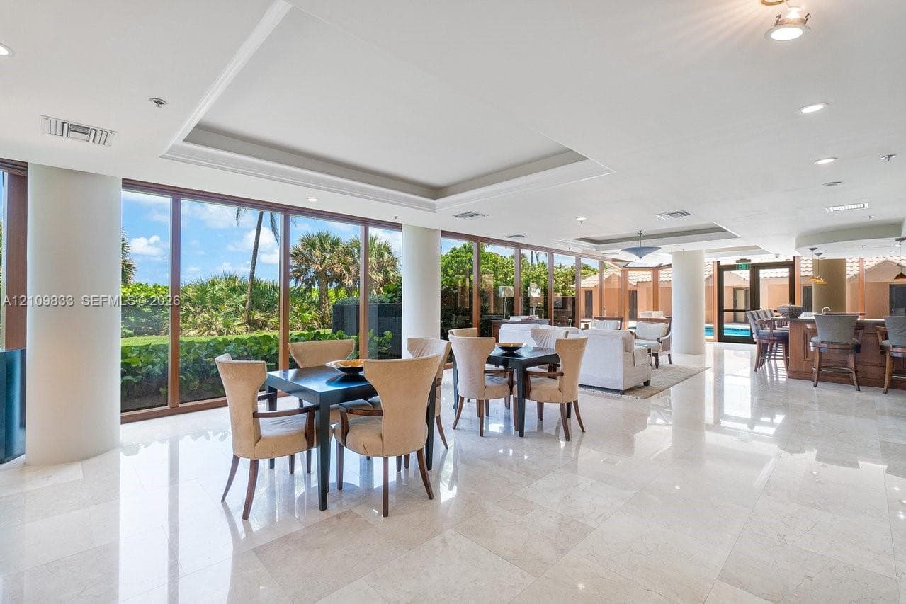 Open-concept interior featuring polished stone flooring, recessed tray ceilings, floor-to-ceiling windows with wood-finish frames, and a built-in wet bar