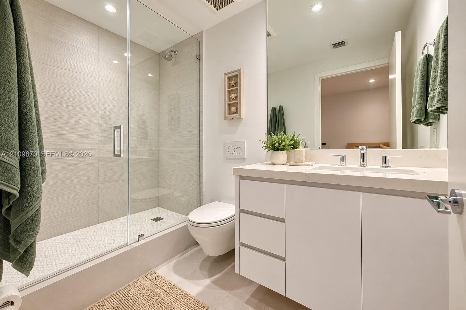 Bathroom featuring a white vanity with an integrated countertop, wall-mounted toilet, walk-in shower with a glass enclosure, and light-toned tile flooring