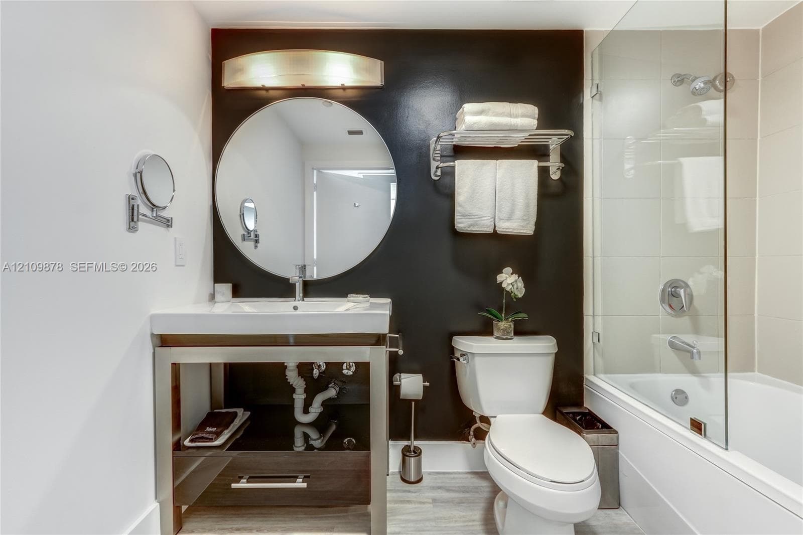 Metal-framed vanity with an integrated rectangular sink, wall-mounted circular mirror, contemporary light fixture, and a bathtub with a glass shower enclosure and tiled surround