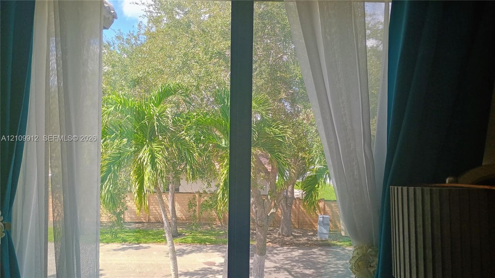 Large glass sliding door framed by a solid vertical mullion, opening toward a fenced backyard with mature palm trees and established landscaping