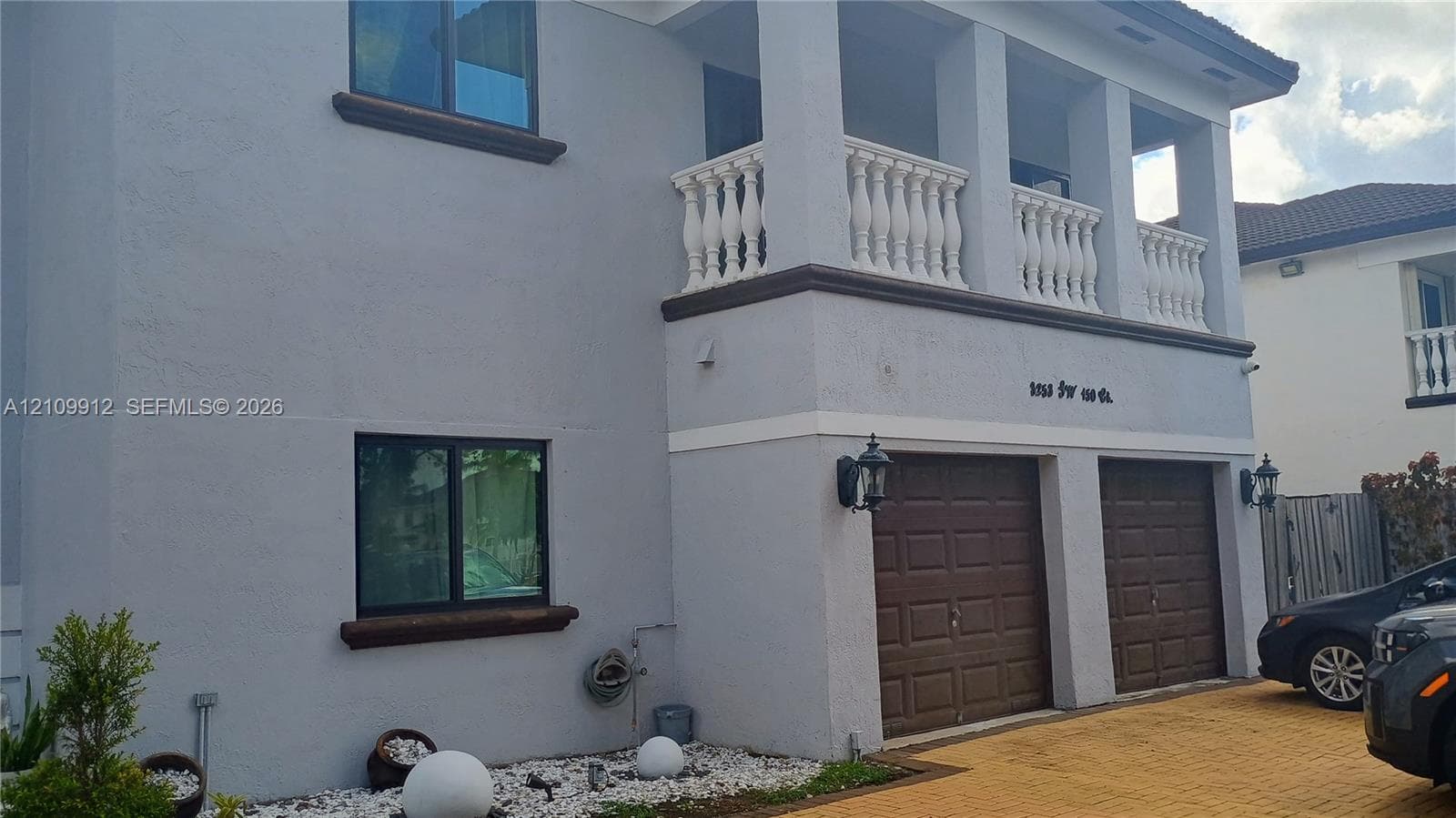 Two-story exterior featuring a stucco facade, covered upper-level balcony with white balustrade railings, two-car garage with paneled doors, and paved driveway