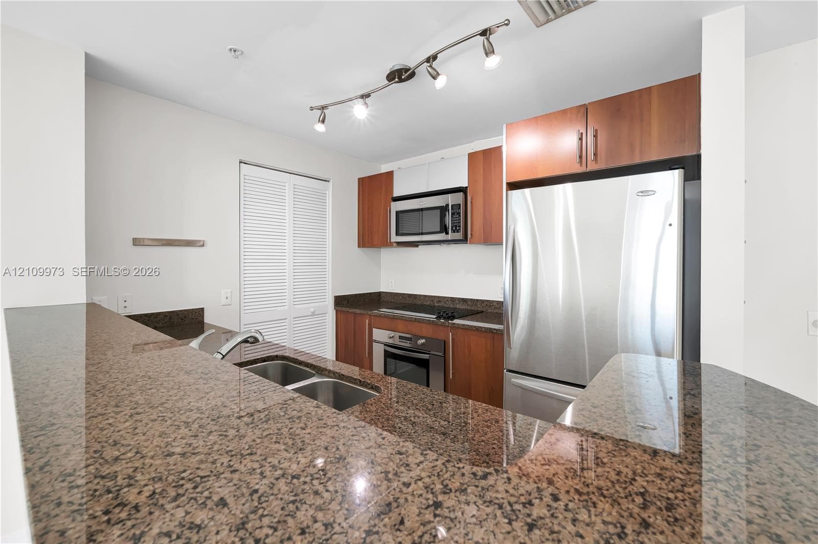 Kitchen featuring polished stone countertops, wood-finish cabinetry, stainless steel appliances, double-basin undermount sink, and track lighting