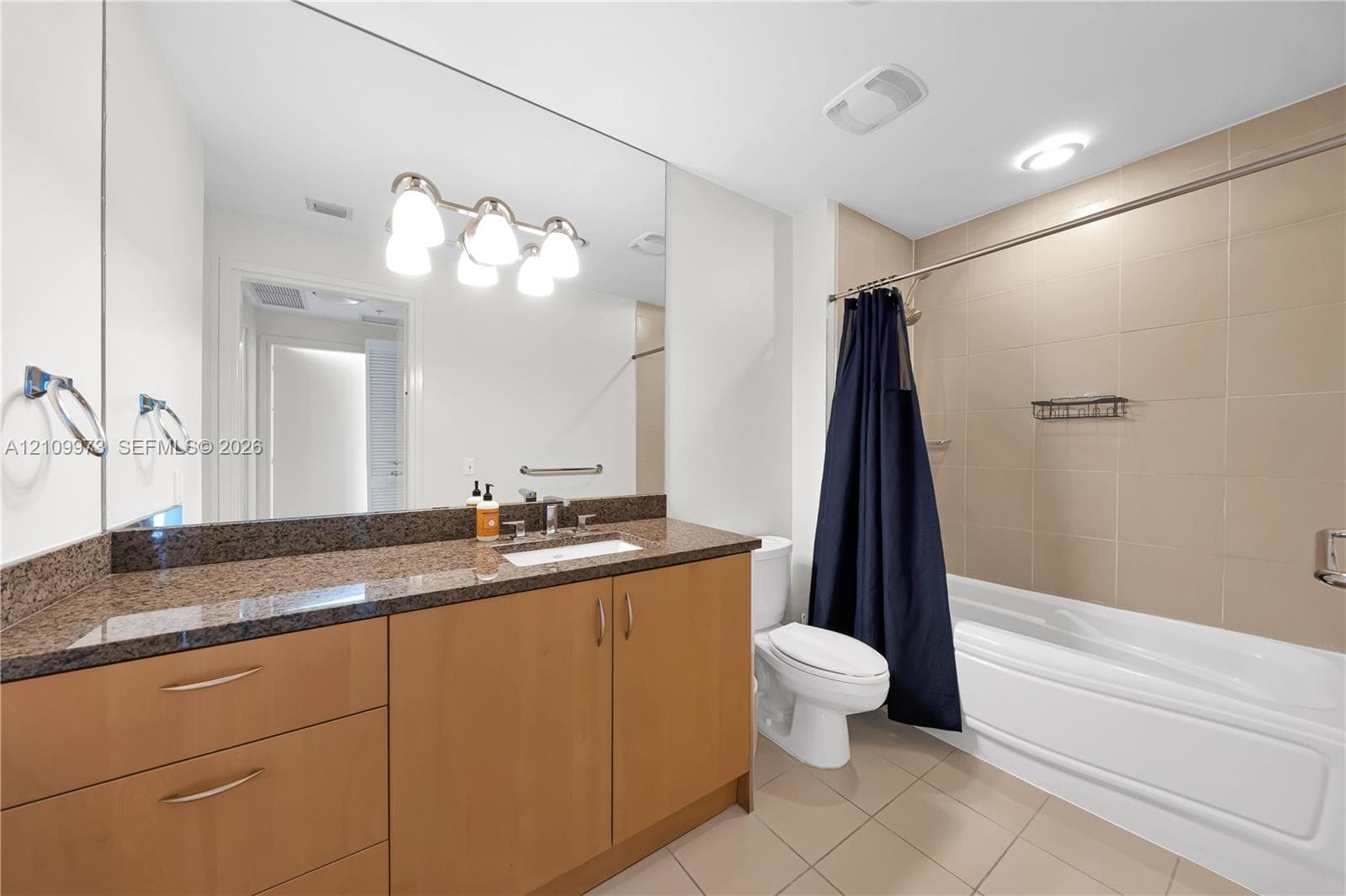 Bathroom featuring light wood cabinetry, stone-finish countertop with an integrated basin, large wall-mounted mirror, and a tub-shower combination with floor-to-ceiling tile surround