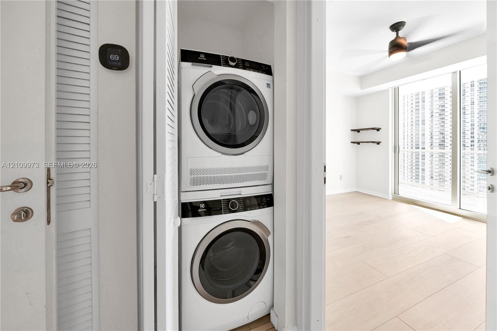 Stackable laundry unit featuring an Electrolux washer and dryer, digital wall-mounted thermostat, wood-finish flooring, ceiling fan with integrated lighting, and wall-mounted shelving