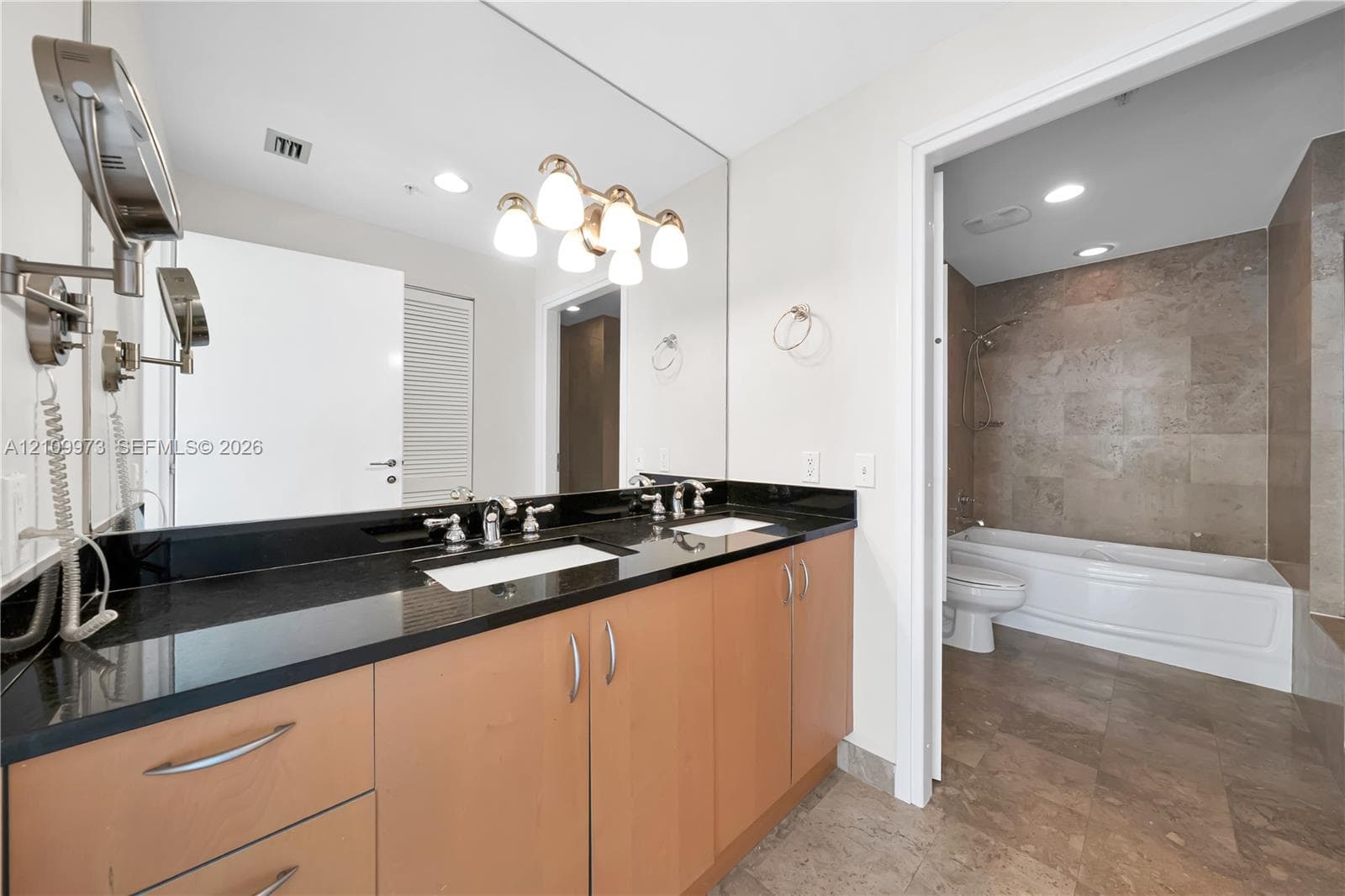 Dual-sink vanity with dark stone countertop, wood-finish cabinetry, full-width mirror, and multi-bulb vanity lighting, featuring a tile-surround bathtub