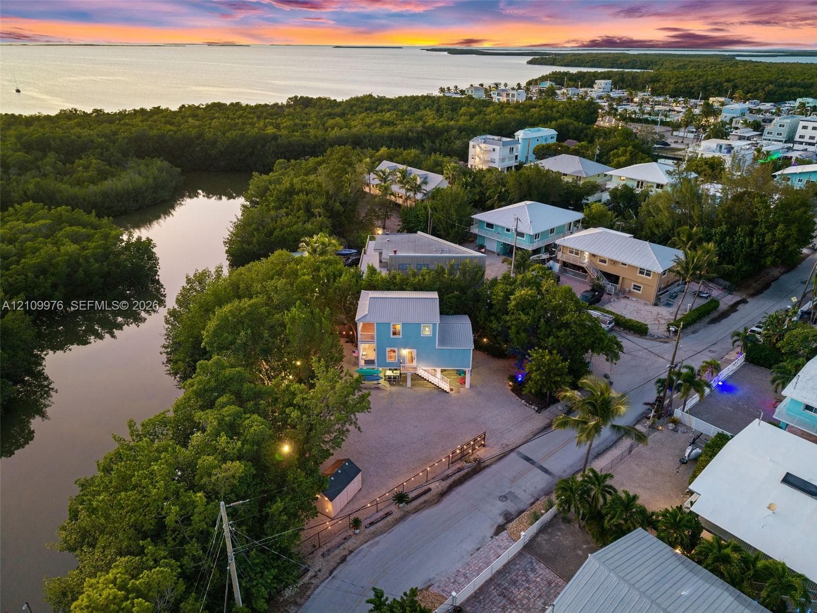 Aerial perspective of a multi-story residence featuring a metal roof, elevated structural design, expansive gravel grounds, and direct positioning along a natural waterway