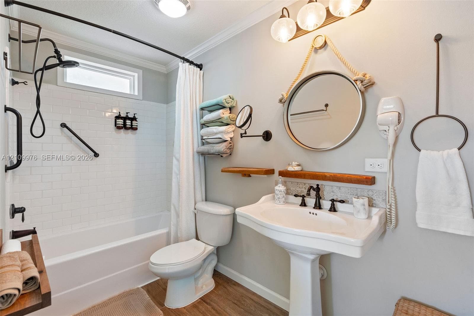 Bathroom featuring white subway tile tub surround, pedestal sink with multi-handle oil-rubbed bronze faucet, round wall-mounted mirror, wood-finish flooring, and industrial-style multi-light fixture