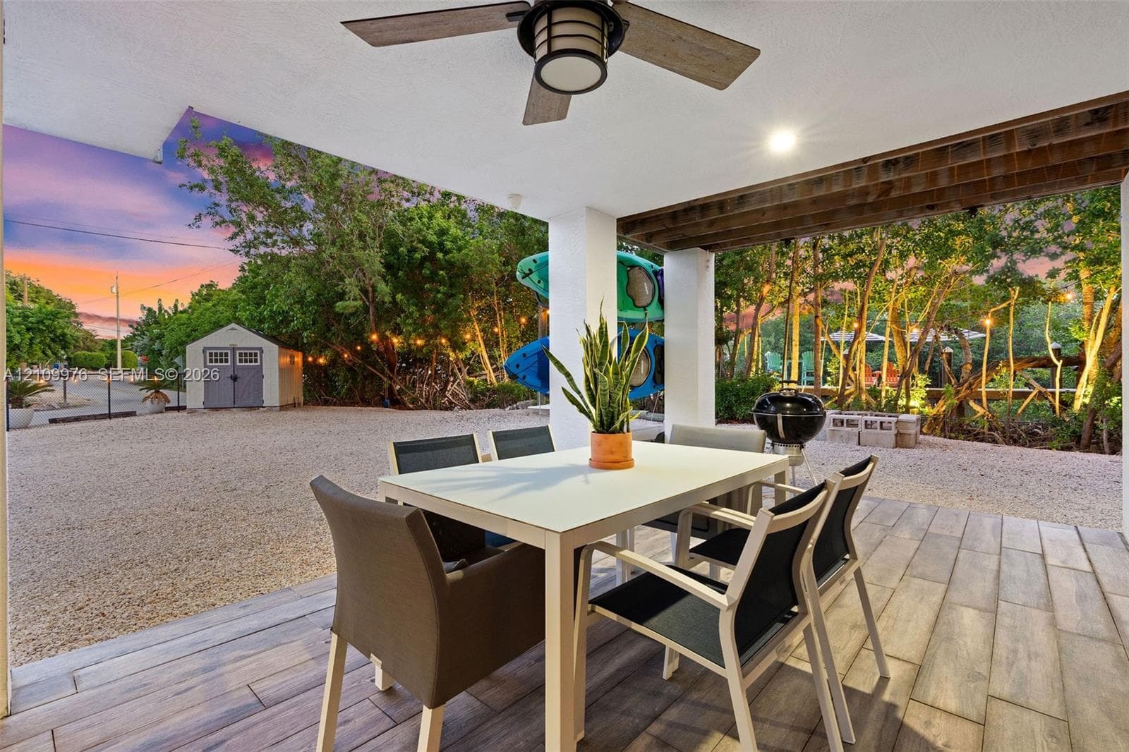 Covered patio featuring wood-finish tile flooring, ceiling fan, and exposed wood-beam detailing, opening to a gravel courtyard with a storage structure