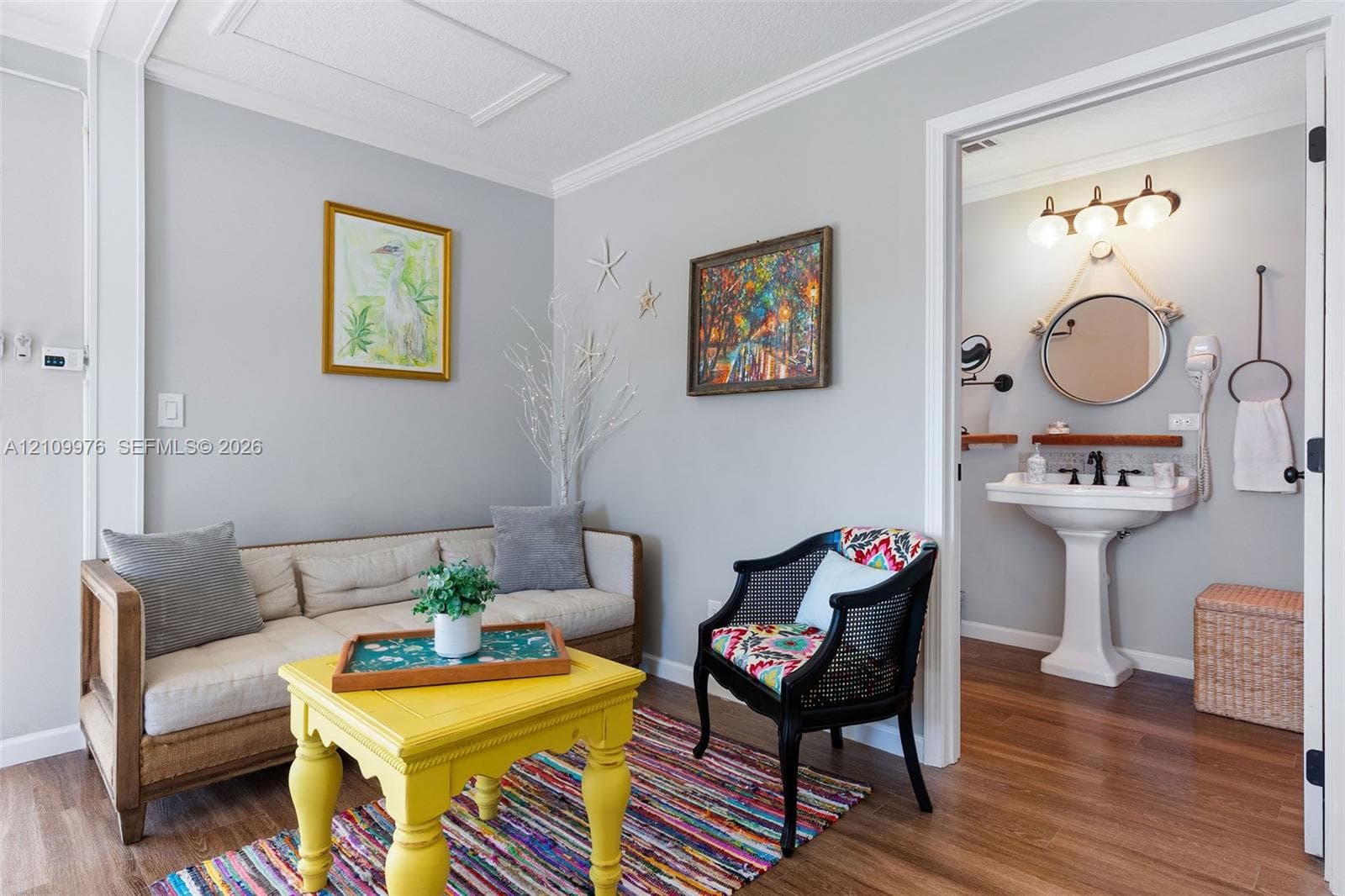 Interior space featuring wood-finish flooring and crown molding, with an opening to a bathroom equipped with a pedestal sink, three-bulb vanity lighting, and a circular mirror suspended by rope