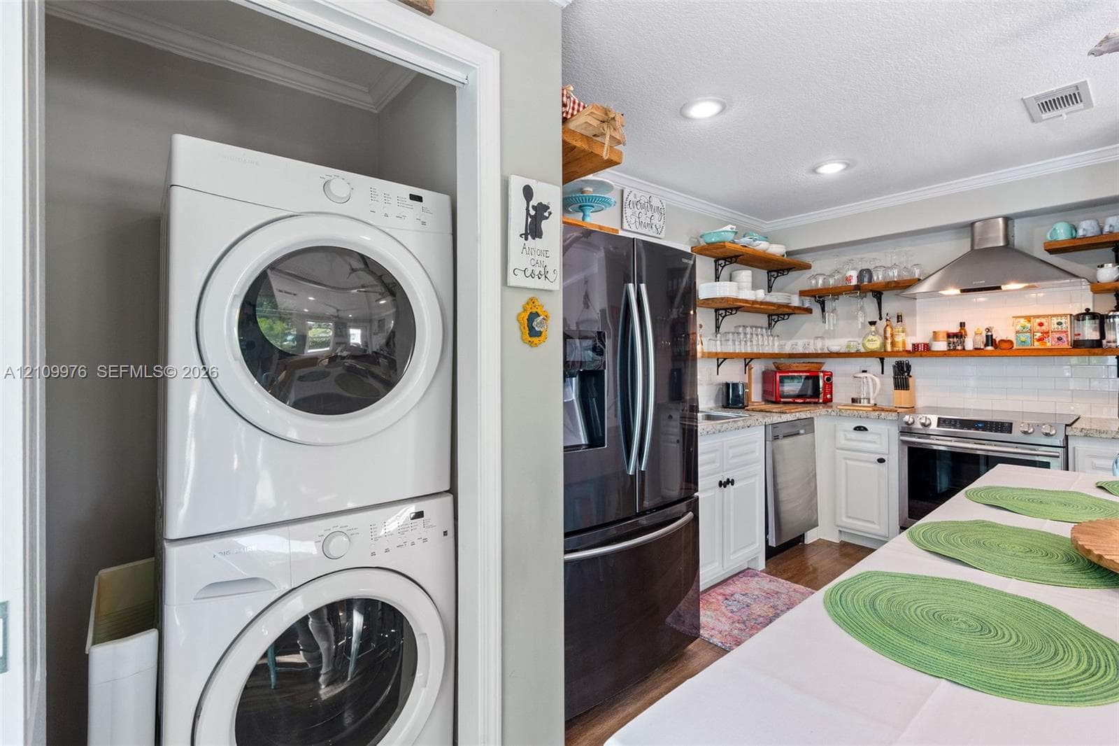 Stacked laundry unit, white cabinetry with stone countertops, stainless steel appliance suite, and open shelving