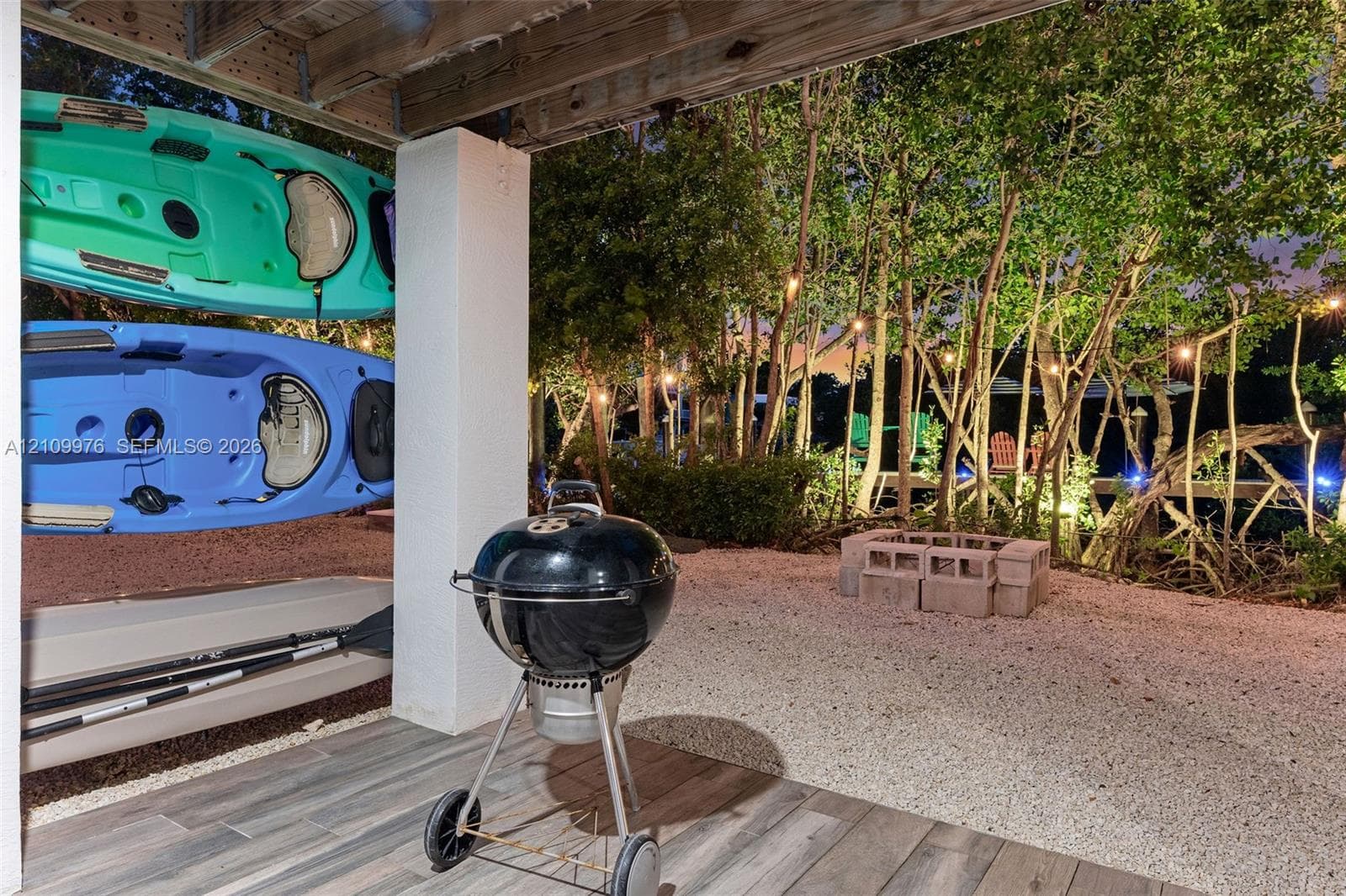 Covered patio with wood-finish tile flooring, crushed stone ground cover, masonry block fire pit, and mature perimeter trees