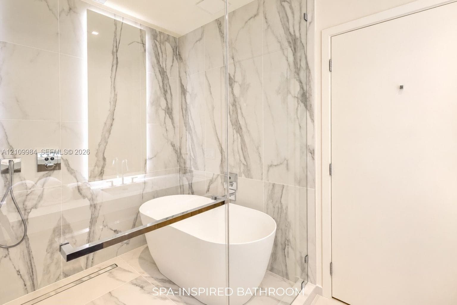Freestanding soaking tub enclosed in a glass-paneled wet room with floor-to-ceiling stone-patterned tile, integrated wall lighting, and chrome fixtures