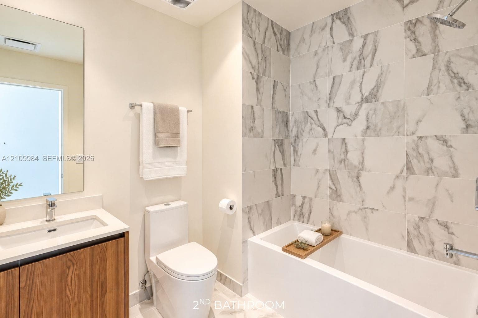 Bathroom featuring a wood-finish vanity with a quartz countertop, rectangular basin, large wall-mounted mirror, and a soaking tub with full-height stone-patterned wall tile