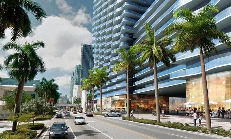 High-rise glass facade with curvilinear balconies, floor-to-ceiling glass exterior, stone-paved pedestrian entrance, and mature palm landscaping
