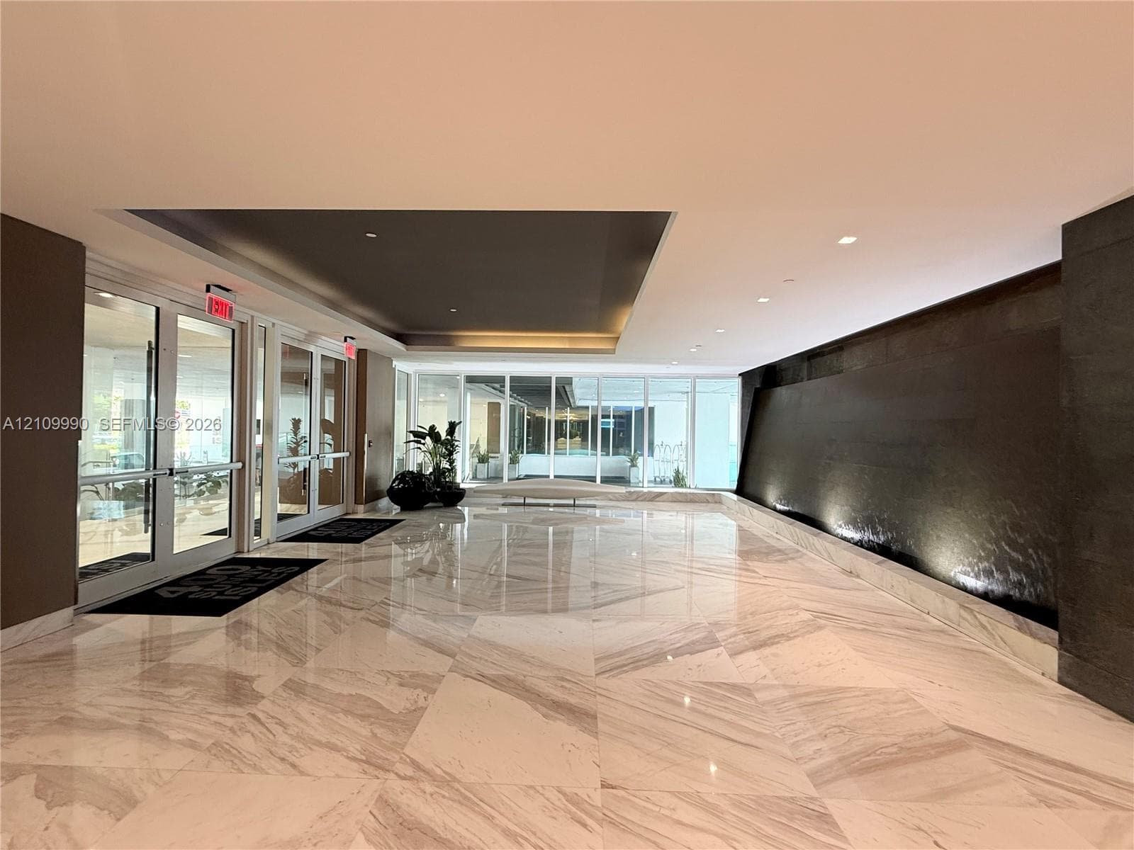 Spacious lobby featuring polished stone flooring in a geometric pattern, an expansive dark-toned accent water wall, recessed ceiling detailing, and glass-paneled entry doors