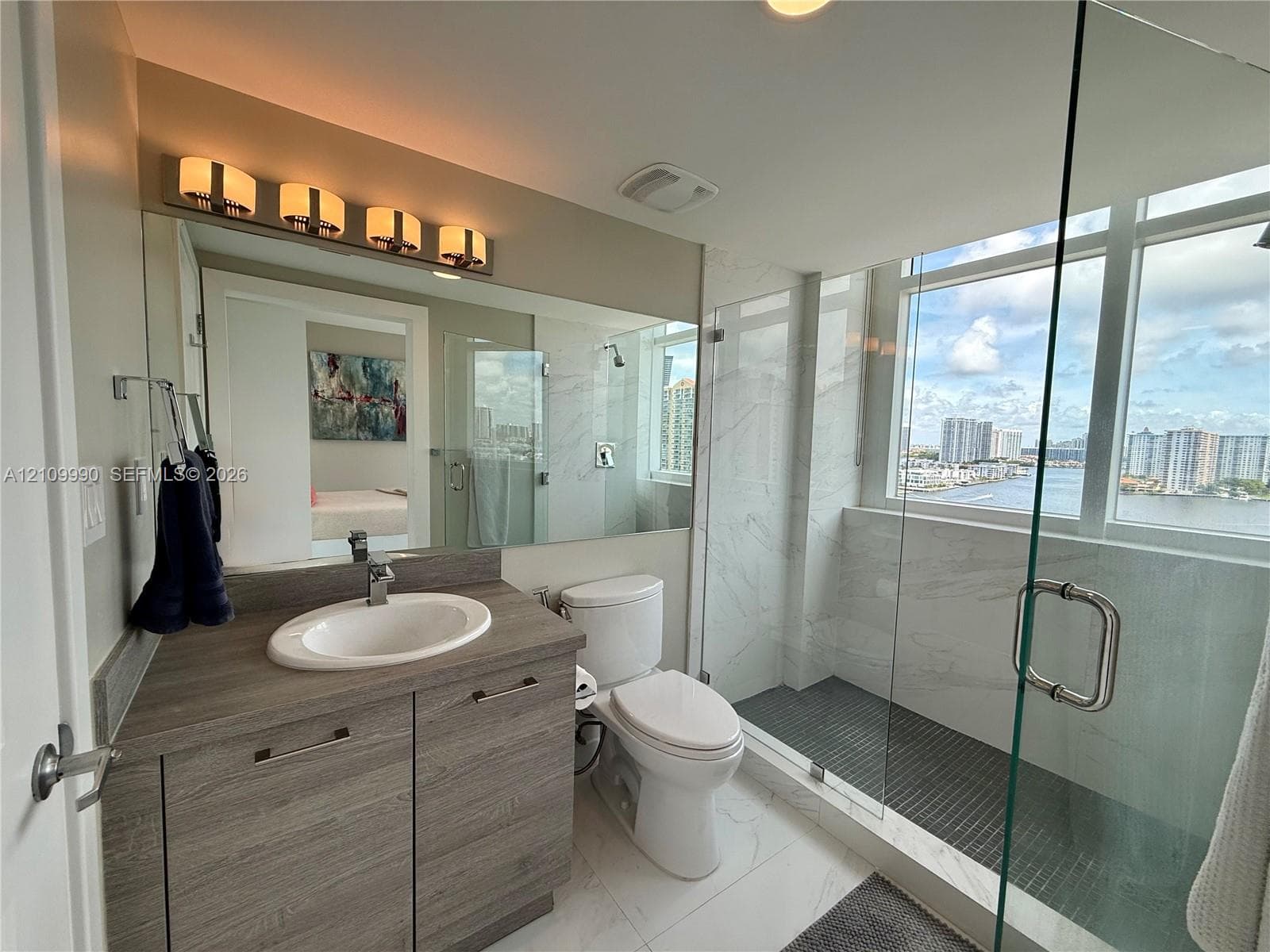 Bathroom featuring a wood-finish vanity with a drop-in sink, large wall-mounted mirror, walk-in shower with stone-style surround and glass enclosure, and large-format flooring