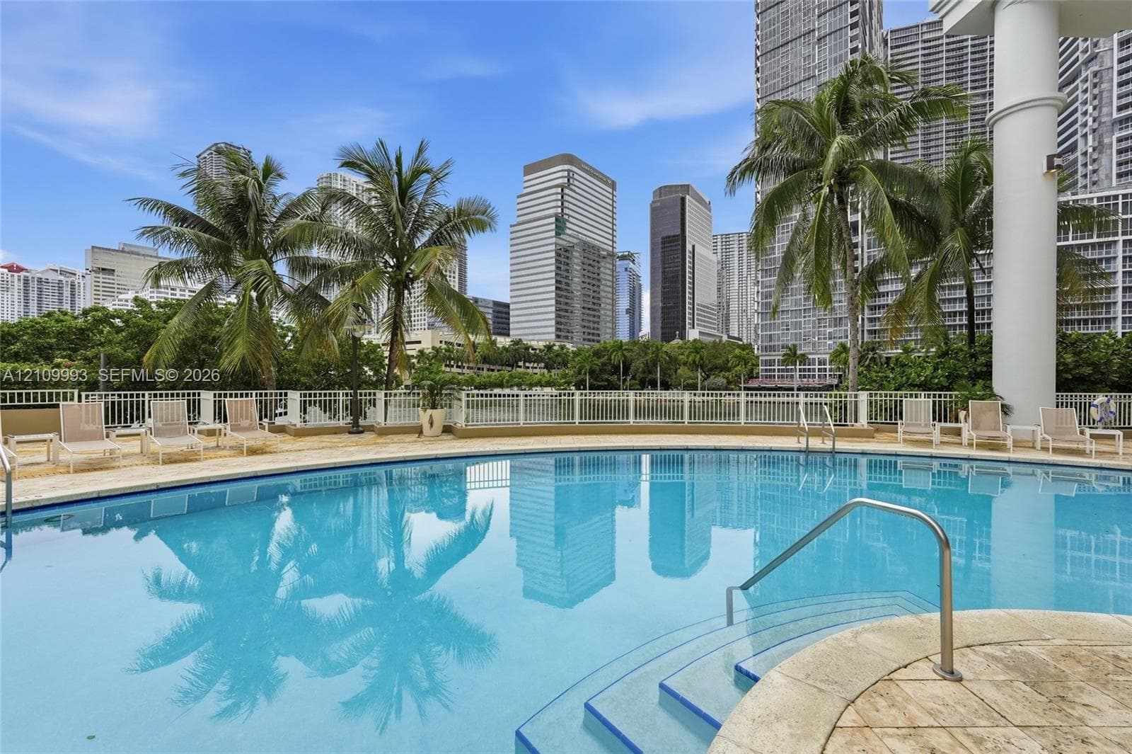 Swimming pool with stone decking, metal handrails, white metal perimeter fencing, and mature palm trees