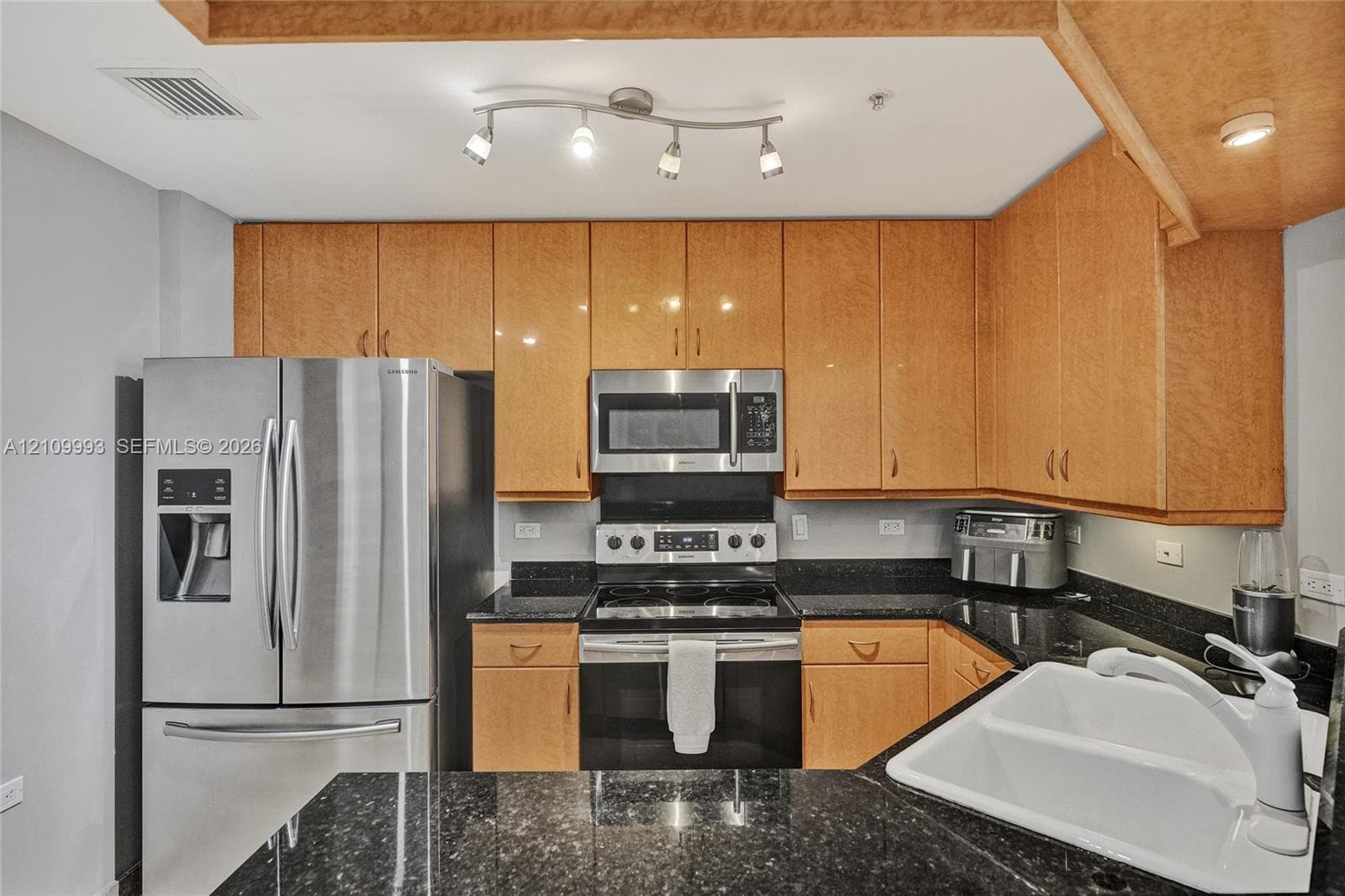 Kitchen featuring flat-panel wood cabinetry, dark stone countertops, a dual-basin white sink with a high-arch faucet, and stainless steel appliances including a side-by-side refrigerator and over-the-range microwave