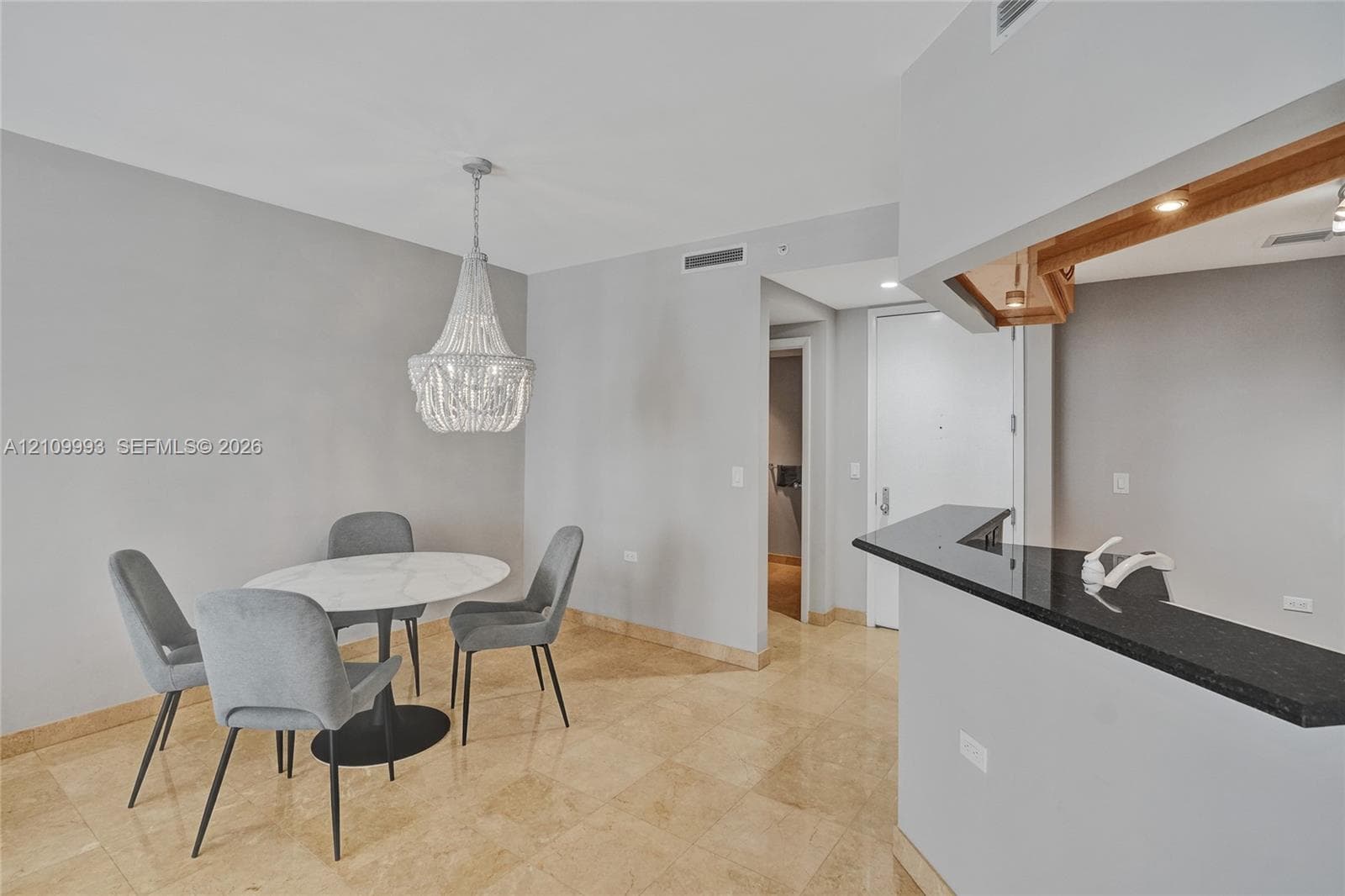 Stone tile flooring, decorative beaded chandelier, and kitchen pass-through with a dark stone countertop