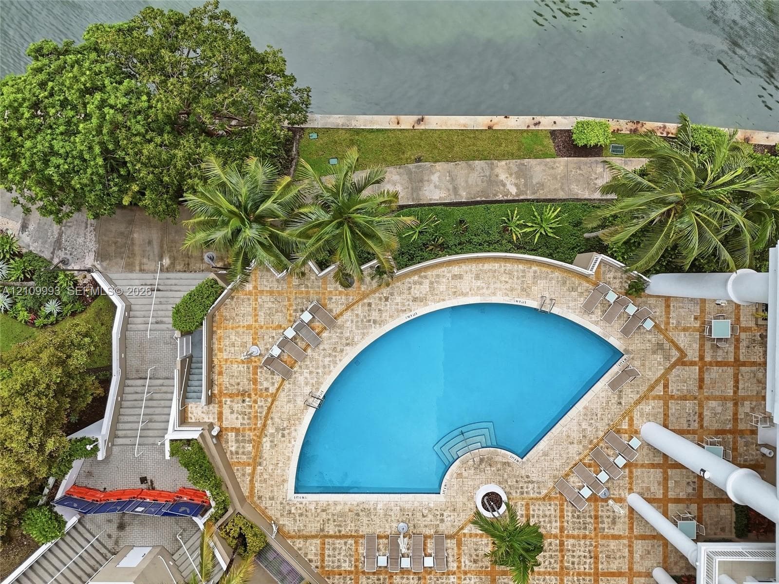 Curved swimming pool with stone deck, tiered landscaping, concrete stairs, and waterfront access