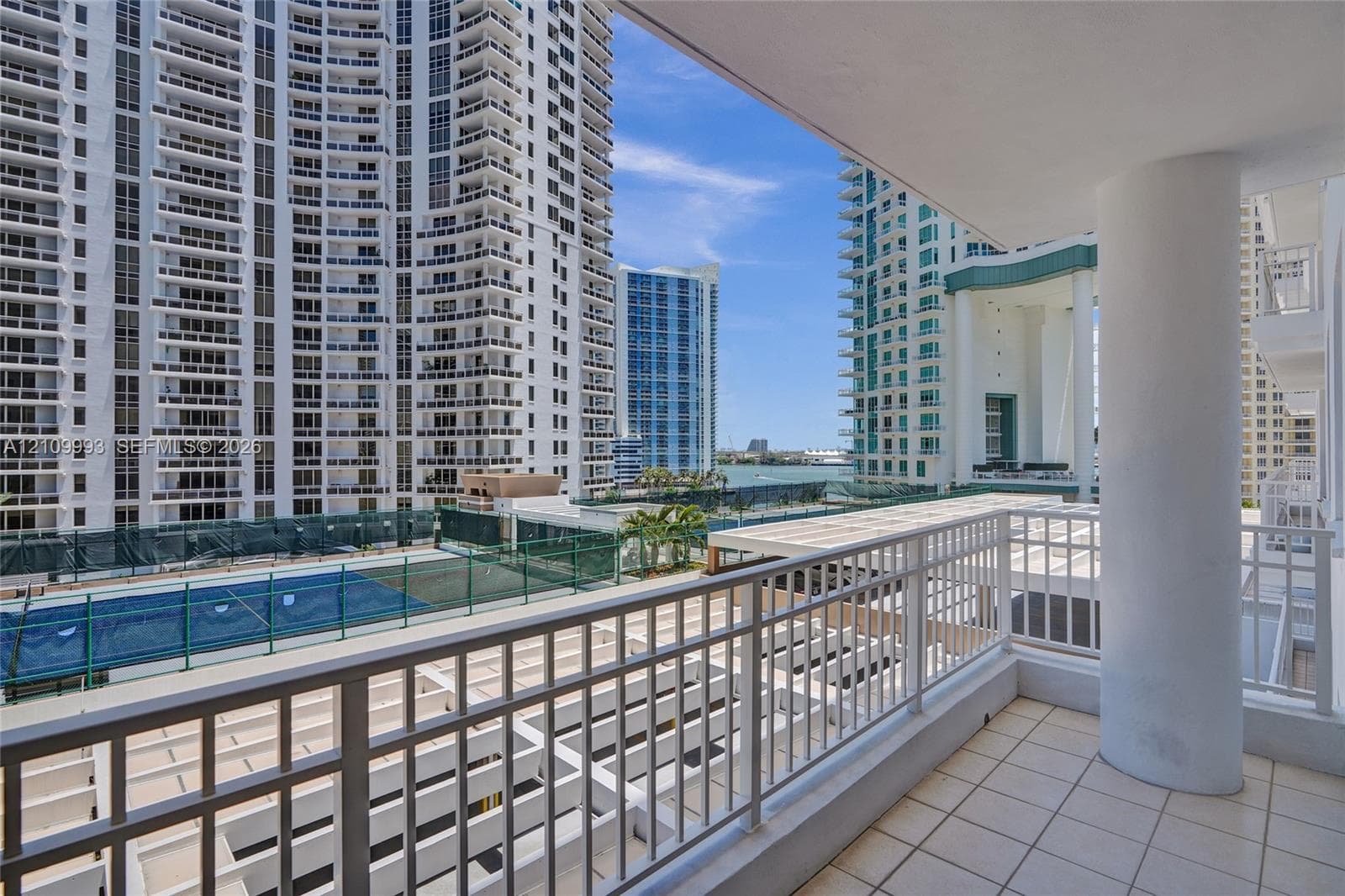 Covered balcony featuring white tile flooring, a painted concrete support column, and metal railings overlooking a tennis court and urban skyline