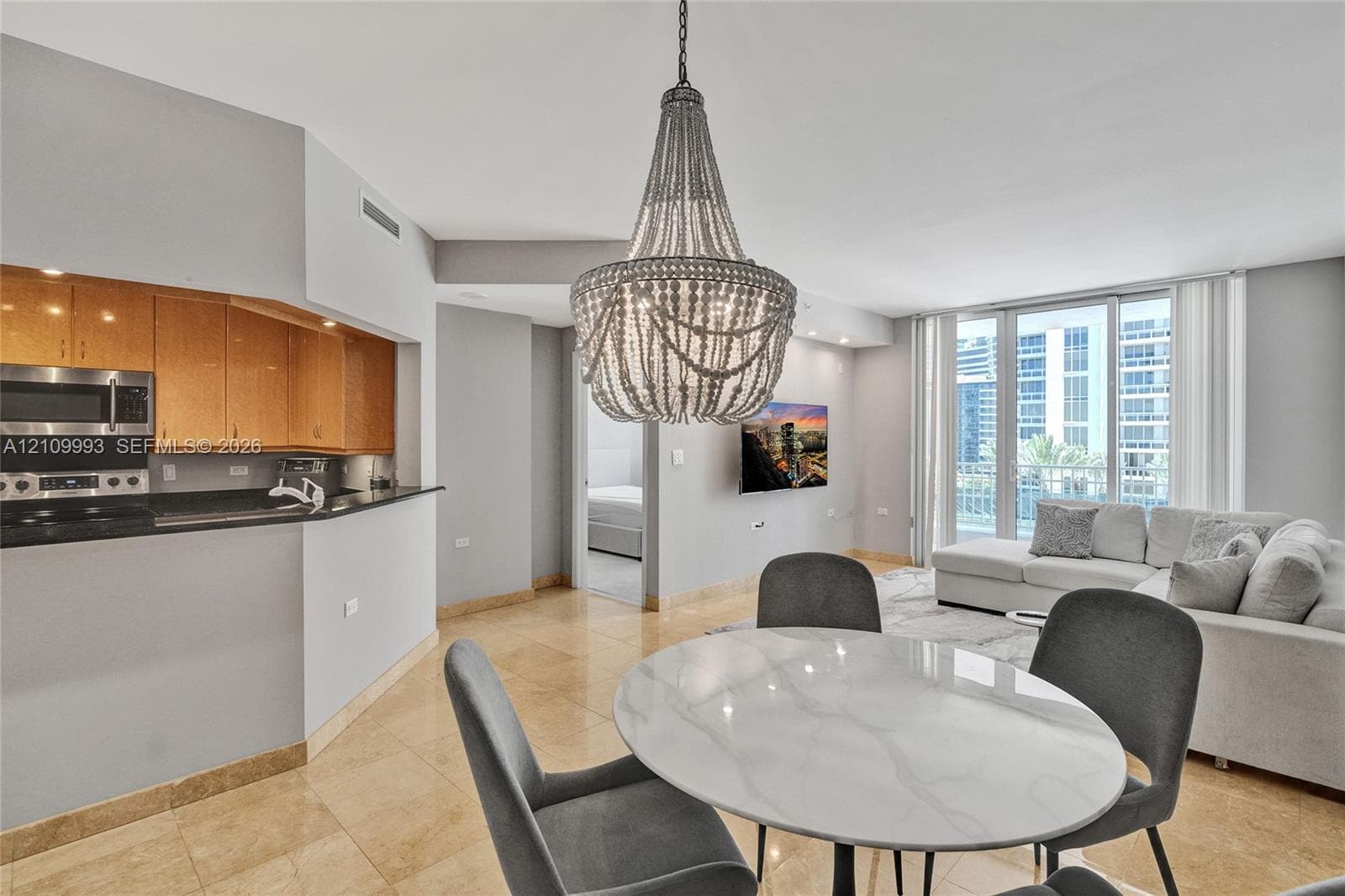 Open-concept kitchen featuring wood-finish cabinetry, black countertops, and stainless steel built-in microwave, complemented by polished stone flooring and a decorative beaded chandelier