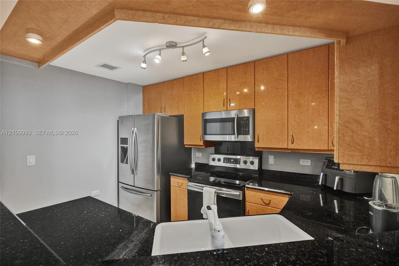 Kitchen featuring high-gloss wood-finish cabinetry, dark solid-surface countertops, a double-basin farmhouse sink, stainless steel appliances, and multi-bulb track lighting