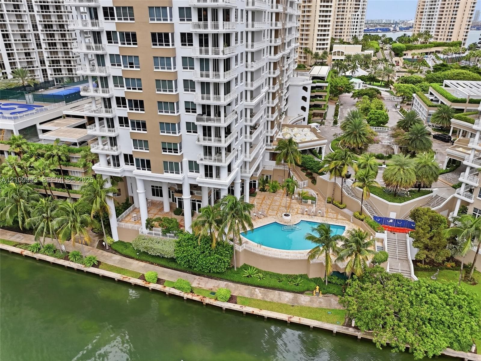 High-rise residential building featuring a swimming pool, waterfront bulkhead, paved terrace, and mature palm landscaping