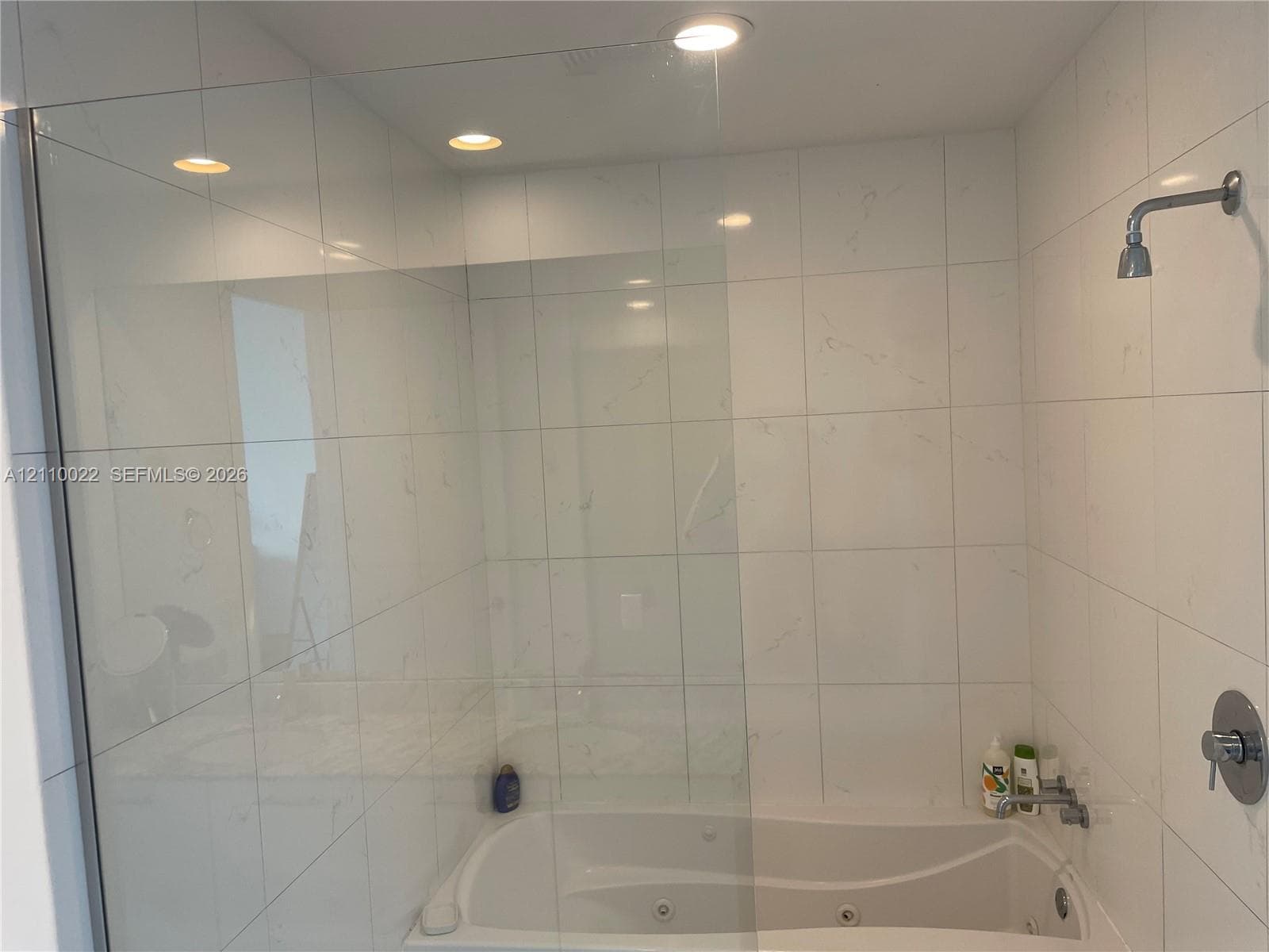 Soaking tub with integrated whirlpool jets, glass shower panel, and white wall tiles featuring subtle veining, complemented by recessed ceiling lighting and chrome fixtures