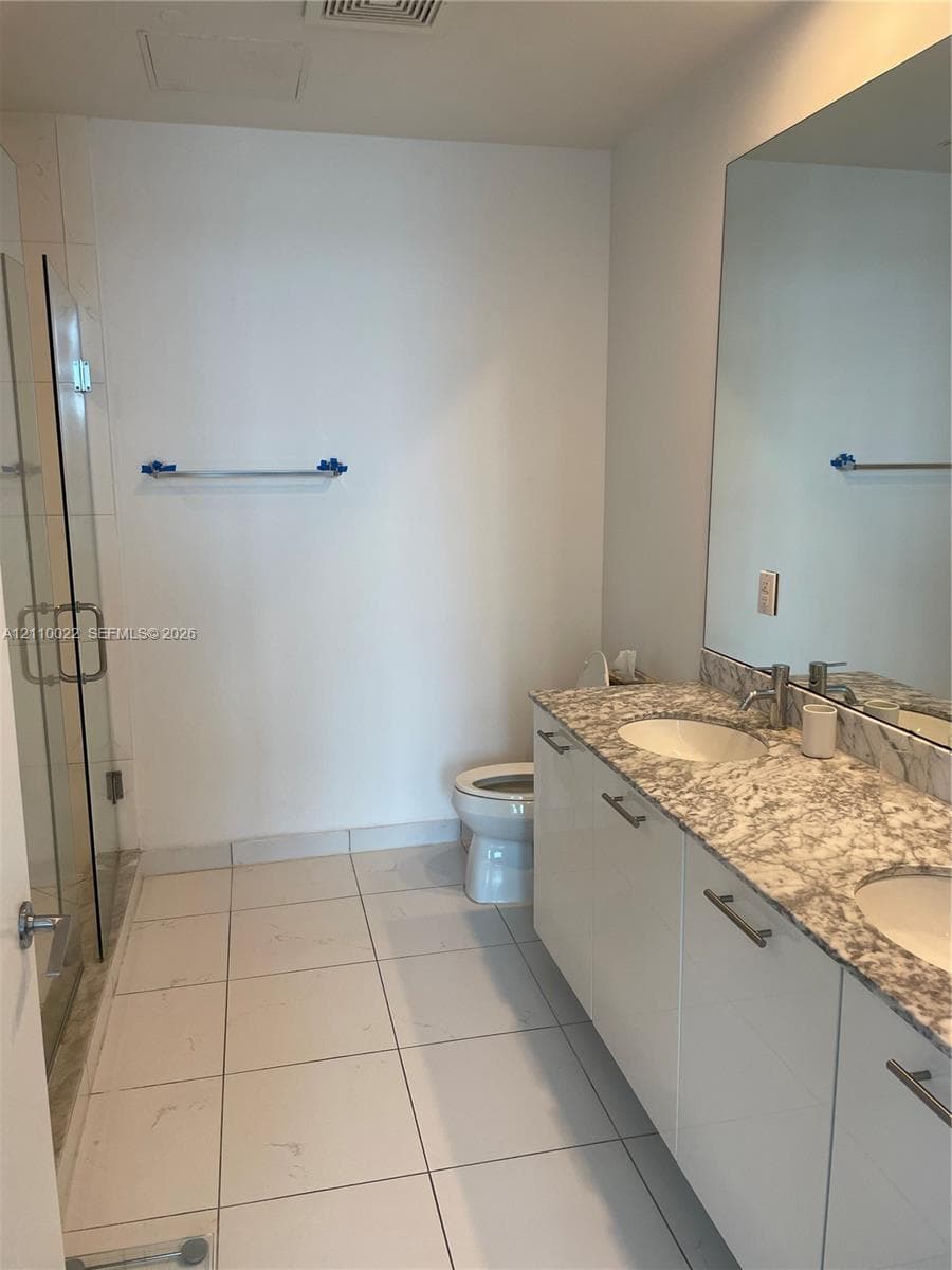 Dual-sink vanity featuring stone-composite countertops, white flat-panel cabinetry, floor-to-ceiling glass-enclosed shower, and large-format ceramic tile flooring